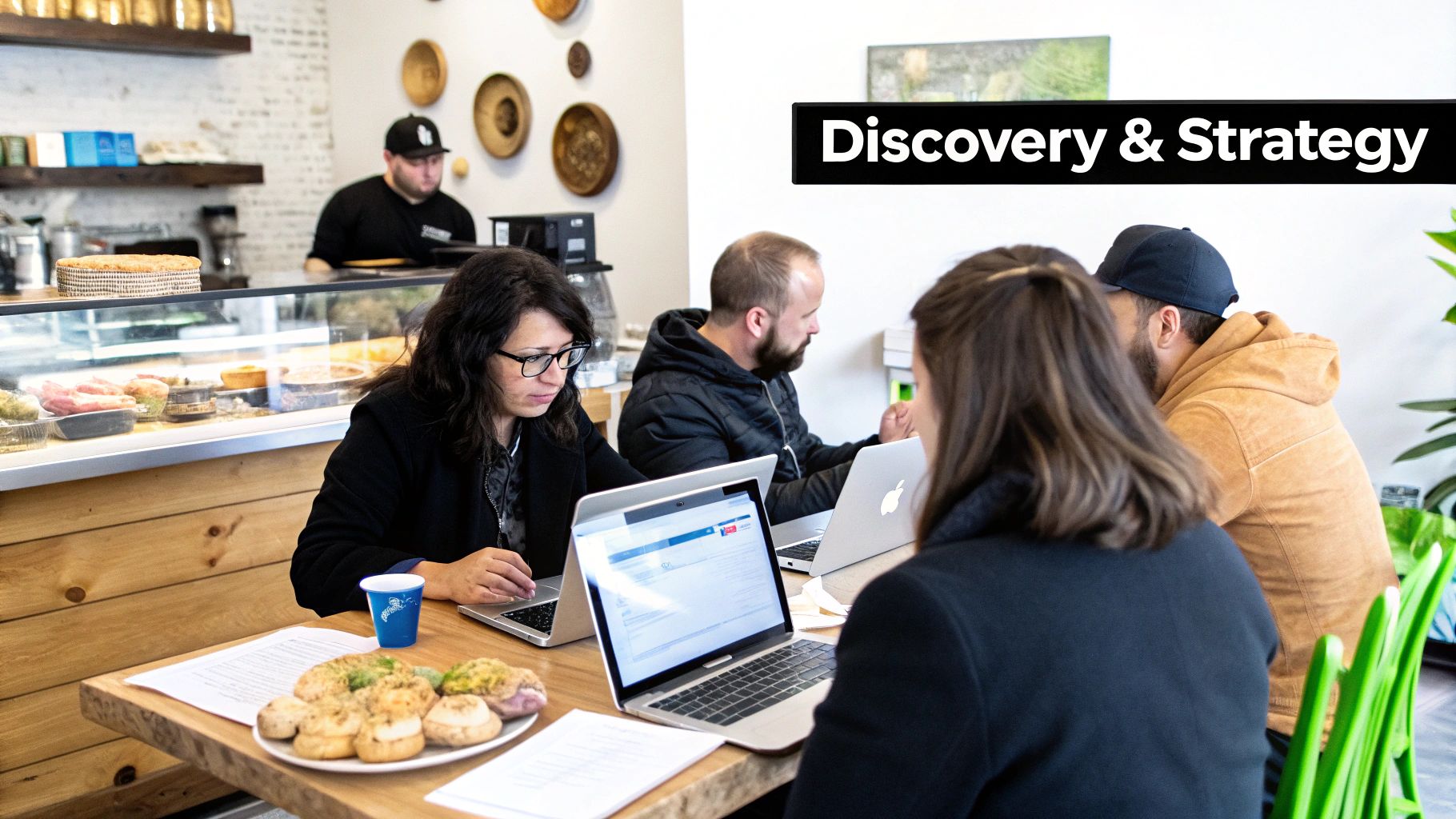 A diverse team of people working on laptops at a cafe table under a 'Discovery & Strategy' sign.
