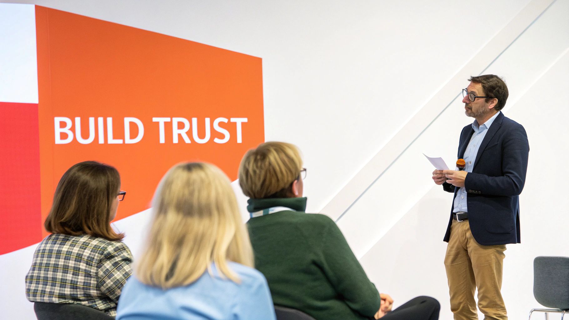 A man in a blazer speaks at a conference, holding a microphone, with "BUILD TRUST" on a screen behind him.