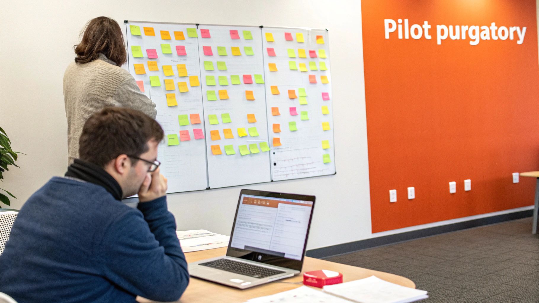 Two people brainstorming in an office, using a whiteboard covered with colorful sticky notes and a laptop.