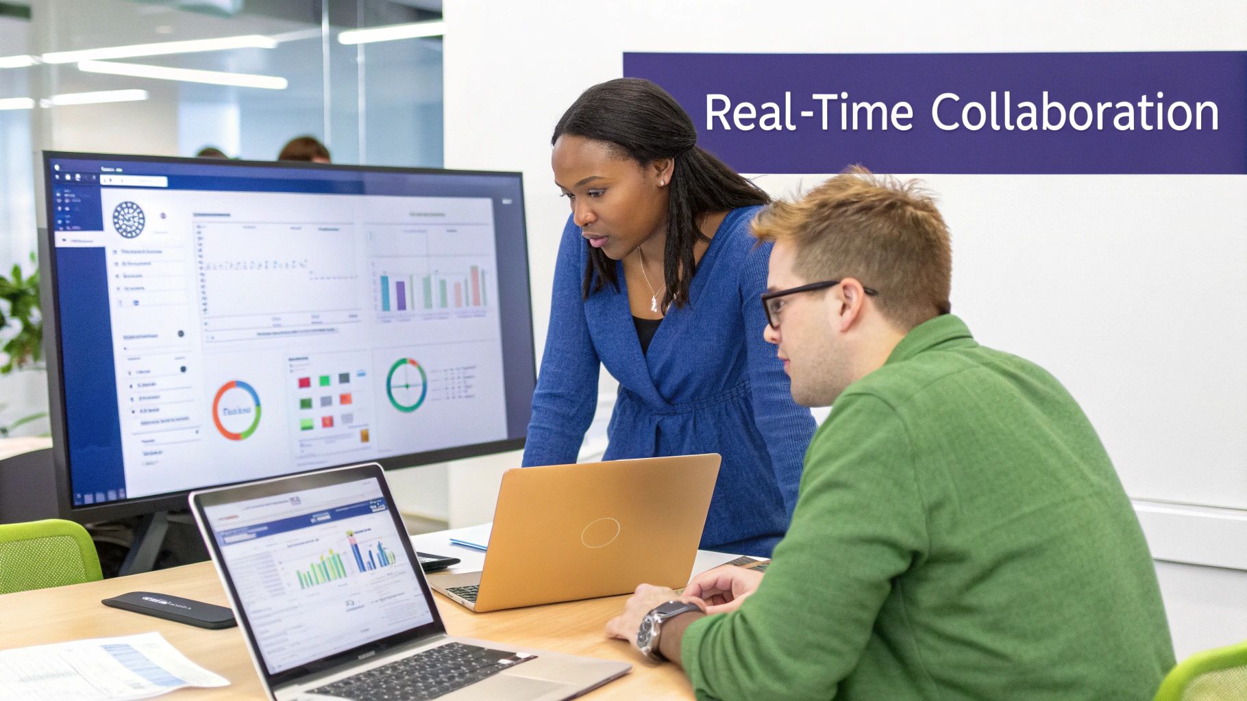 Two colleagues collaborate on data analysis using laptops and a large monitor in an office.