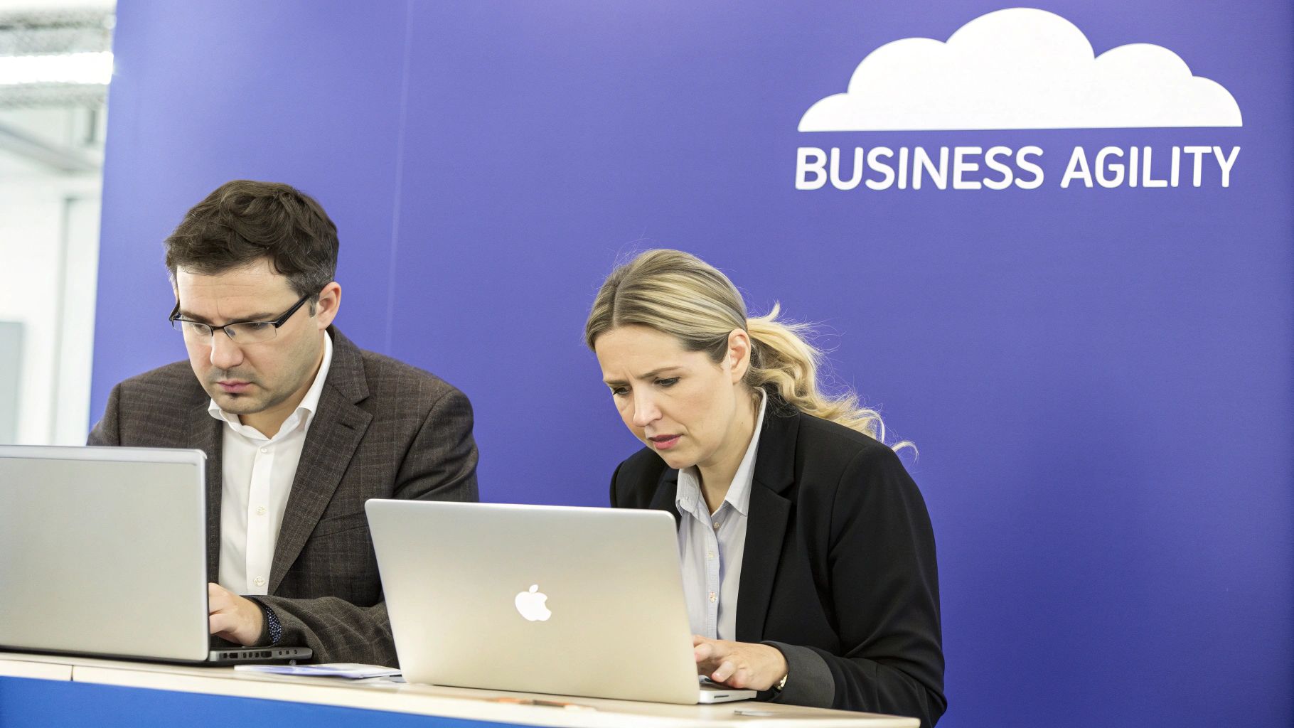 Two focused professionals working on laptops at a business event with a 'BUSINESS AGILITY' sign.