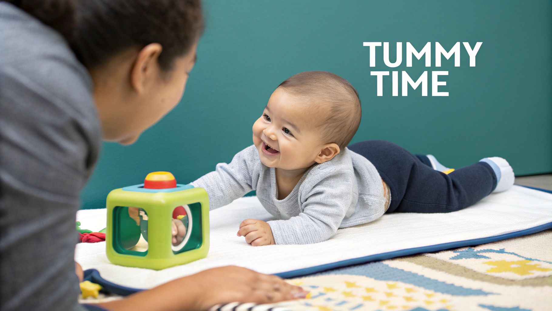 A happy baby smiles brightly during tummy time, looking at an adult with a green toy.