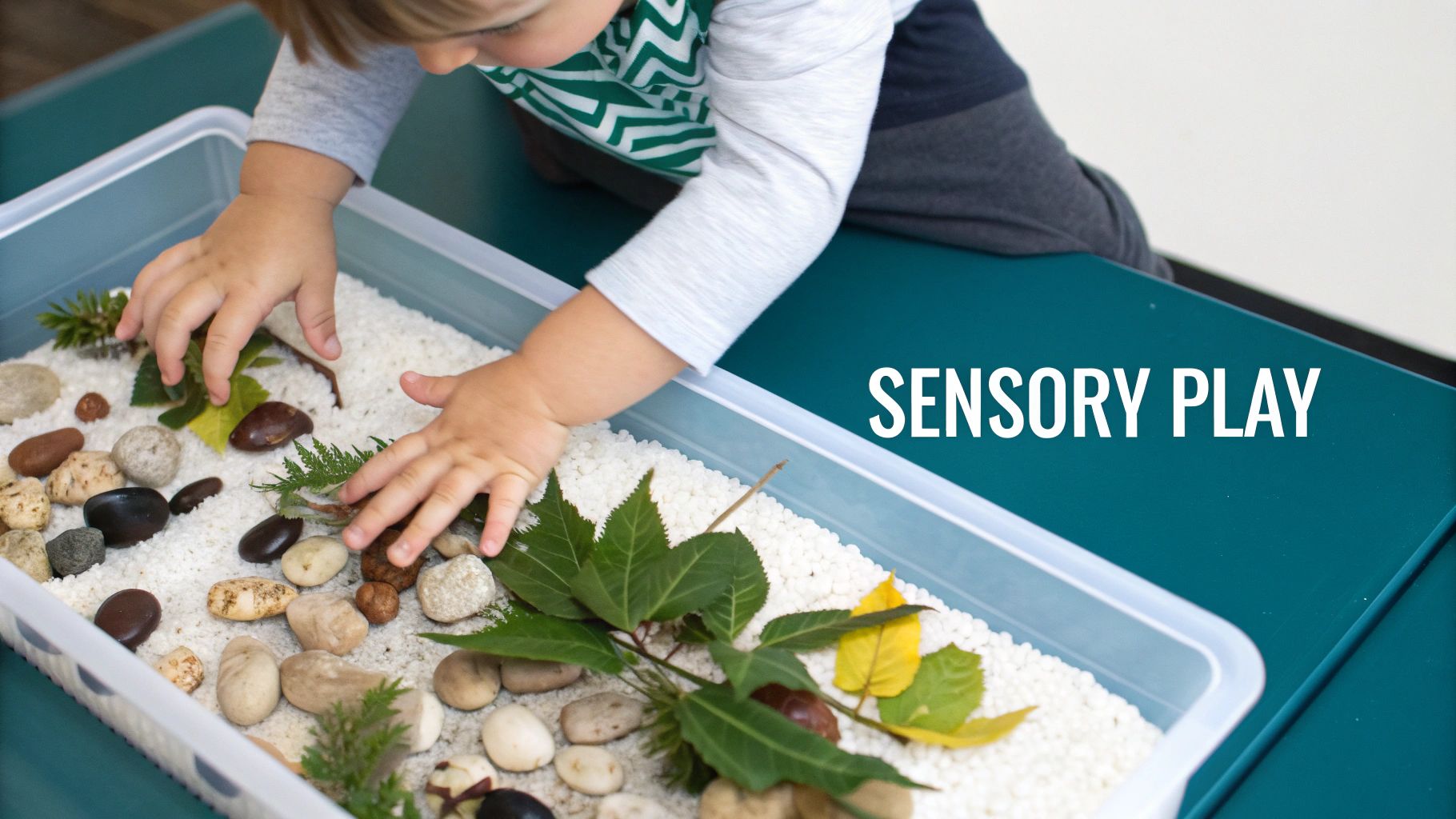A toddler's hands explore a sensory bin filled with white beads, natural rocks, and green leaves.