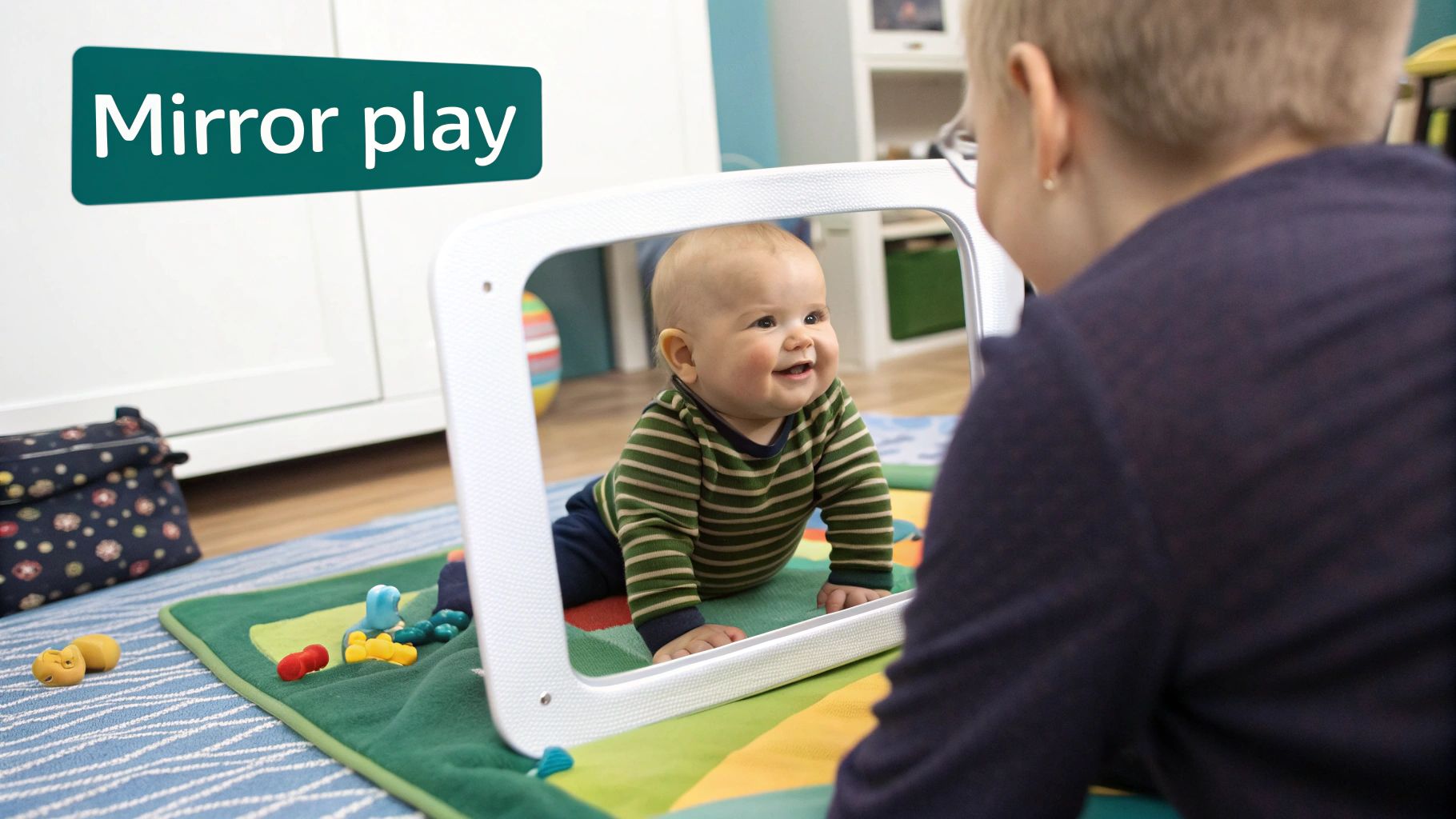 A happy baby on a colorful play mat smiles at its reflection in a mirror, while an adult watches.