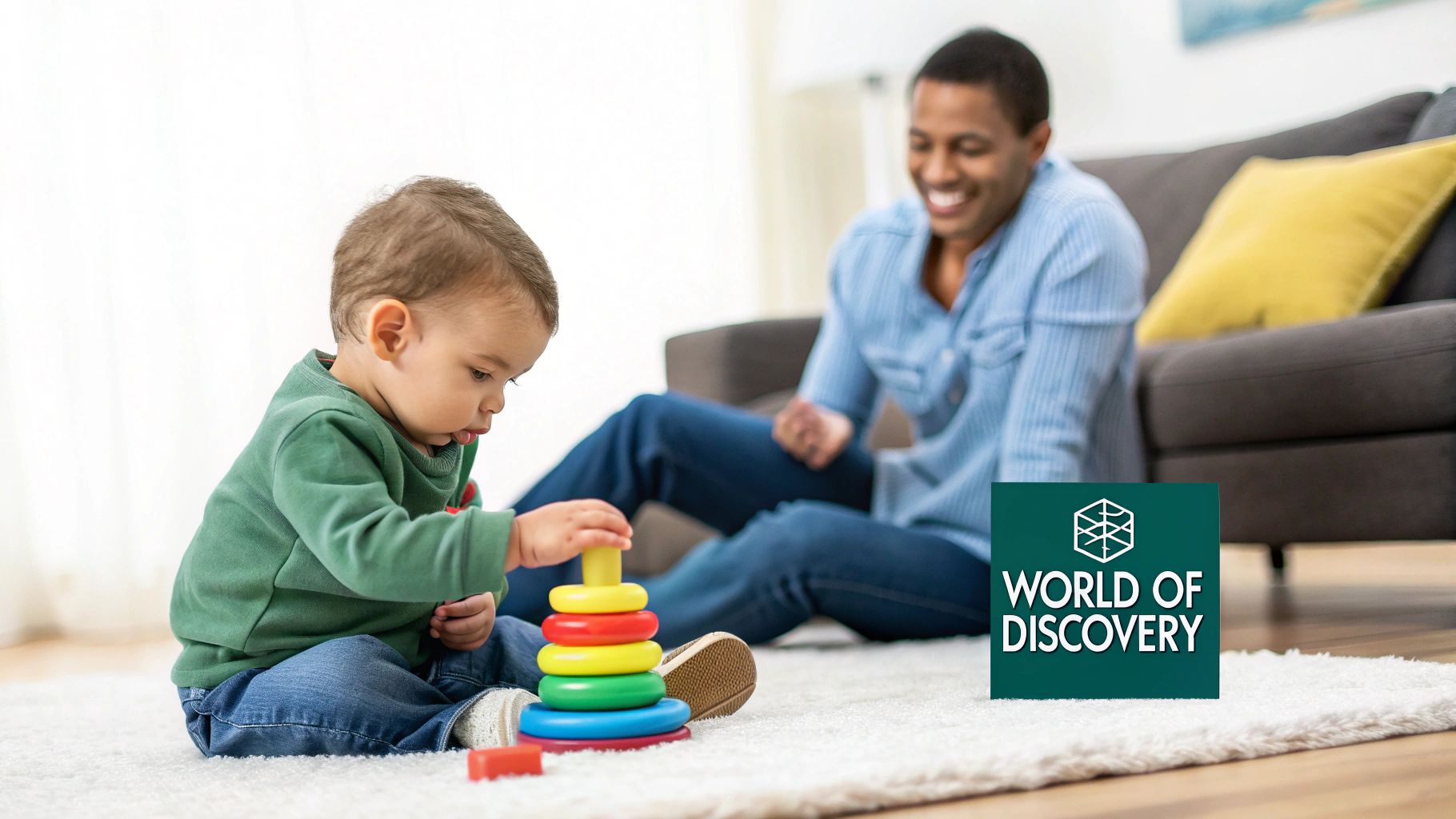 A happy baby plays with a colorful stacking toy on a white rug, with a smiling adult in the background.