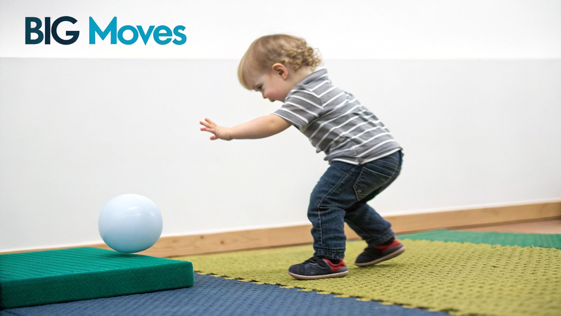 An 18-month-old child plays with a light blue ball on a green mat, learning to balance.