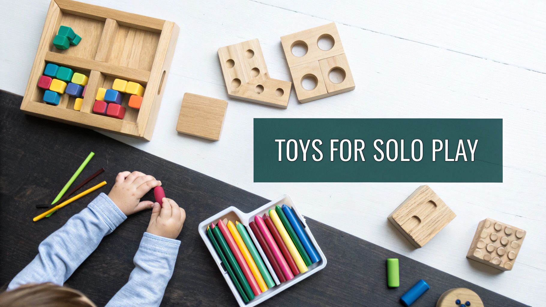 A child's hands play with a red crayon, surrounded by colorful blocks and educational toys on a wooden table.