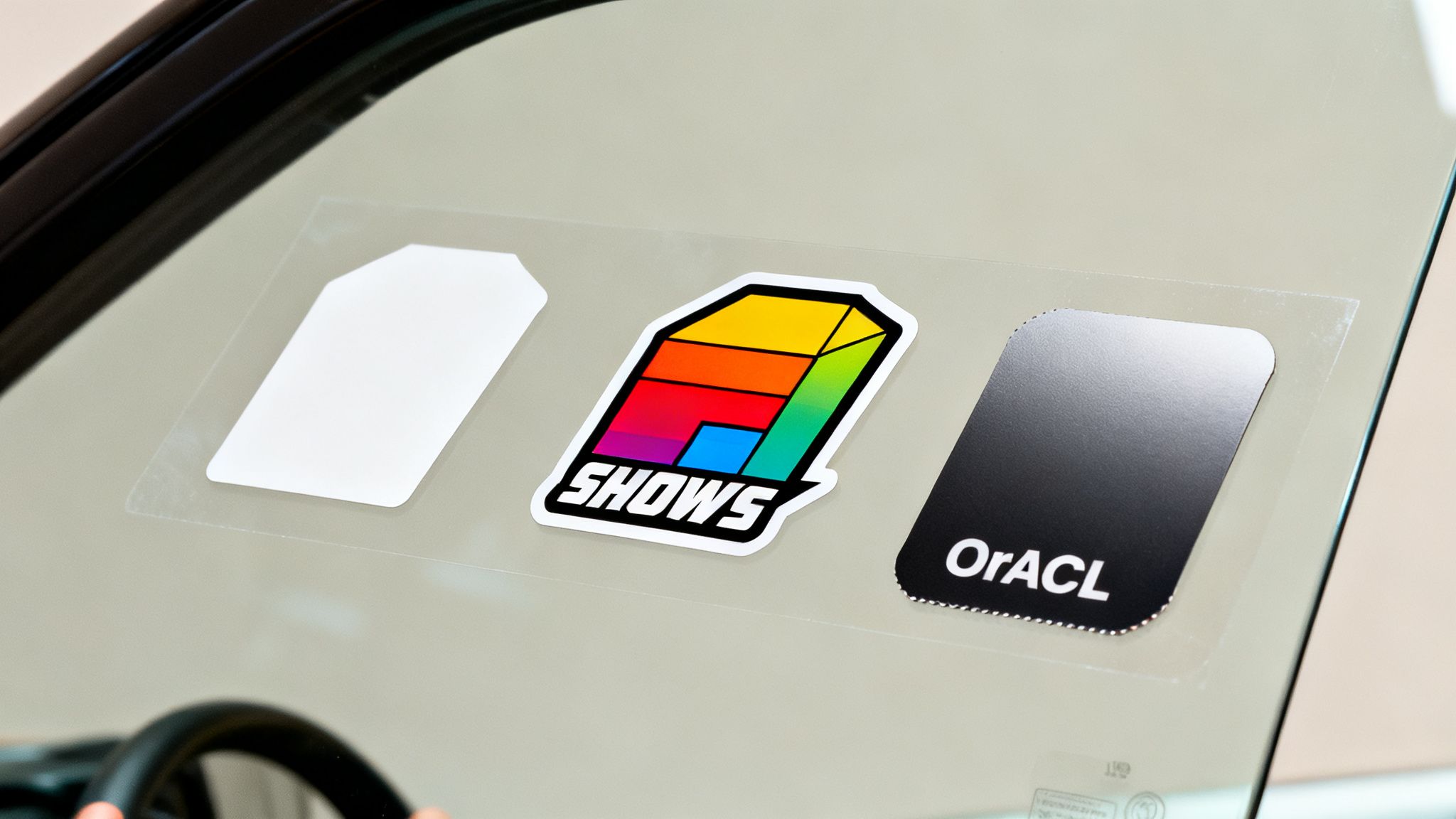 Three diverse decals affixed to a car window, featuring white, colorful 'SHOWS', and black 'OrACL' designs.