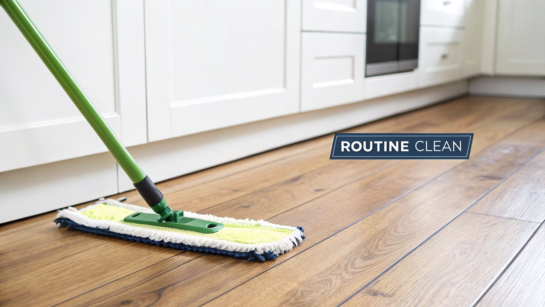 A green flat mop cleaning a light brown wooden floor in a modern kitchen with white cabinets.