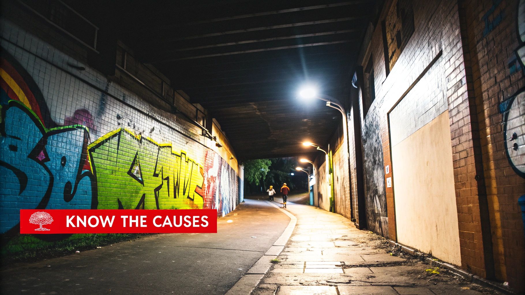 Graffiti-covered tunnel with streetlights illuminating a curved pathway, featuring two individuals walking, emphasizing the importance of strategic lighting and visibility in enhancing property safety and deterring vandalism.