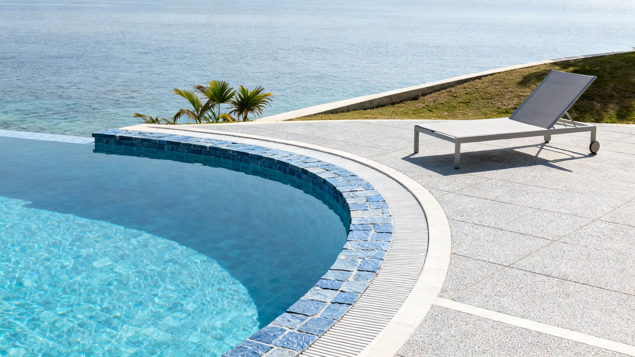 A luxurious curved infinity pool with blue tiles overlooks the ocean, next to a modern lounge chair on a sunlit patio.
