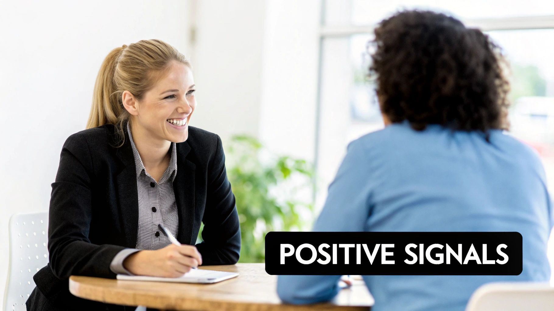 A smiling woman in a black blazer takes notes at a desk, looking engaged during a conversation.