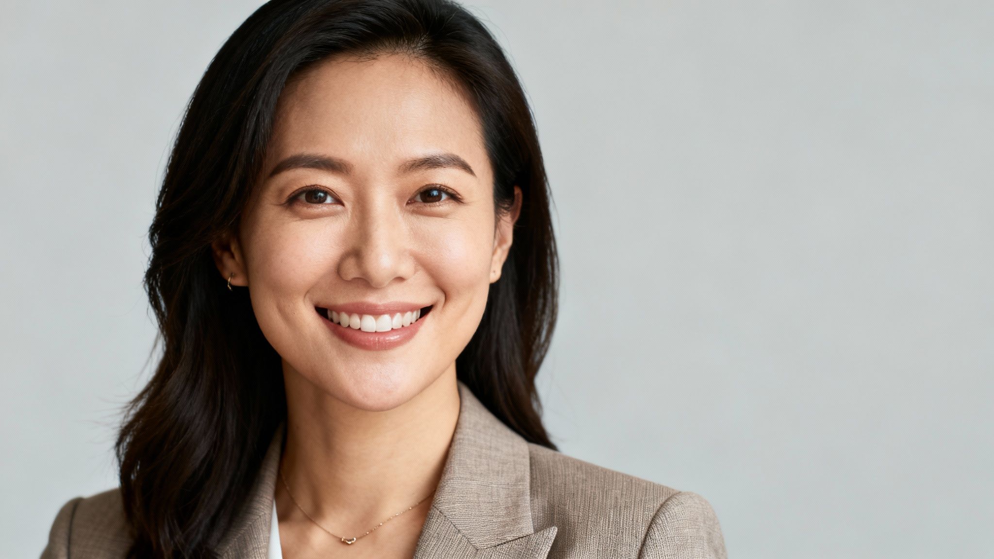 A smiling East Asian woman with long dark hair, wearing a beige blazer and gold necklace.