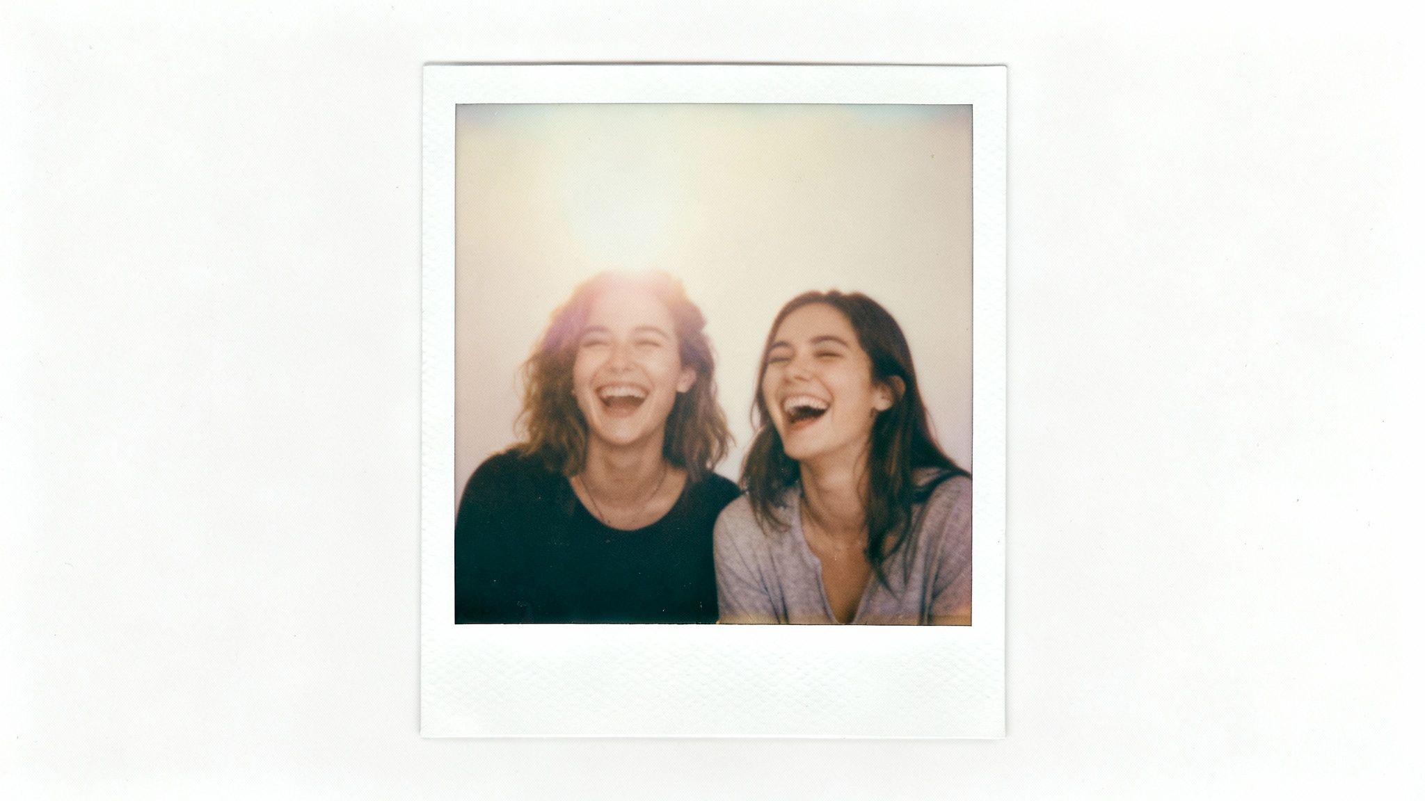 Two joyful young women with curly and straight hair laugh loudly in a vintage Polaroid photo.