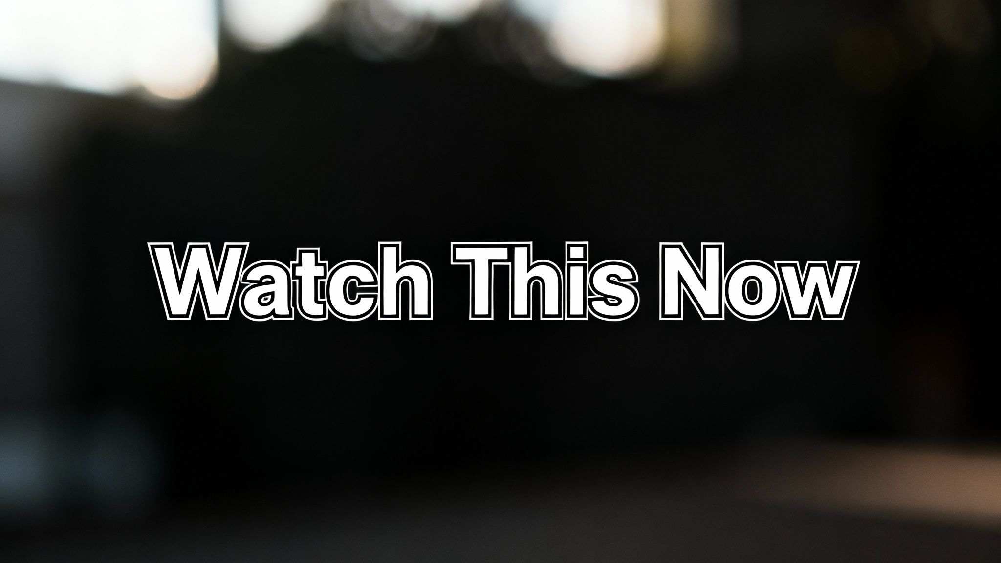 White text 'Watch This Now' with a black outline on a dark, blurry background.