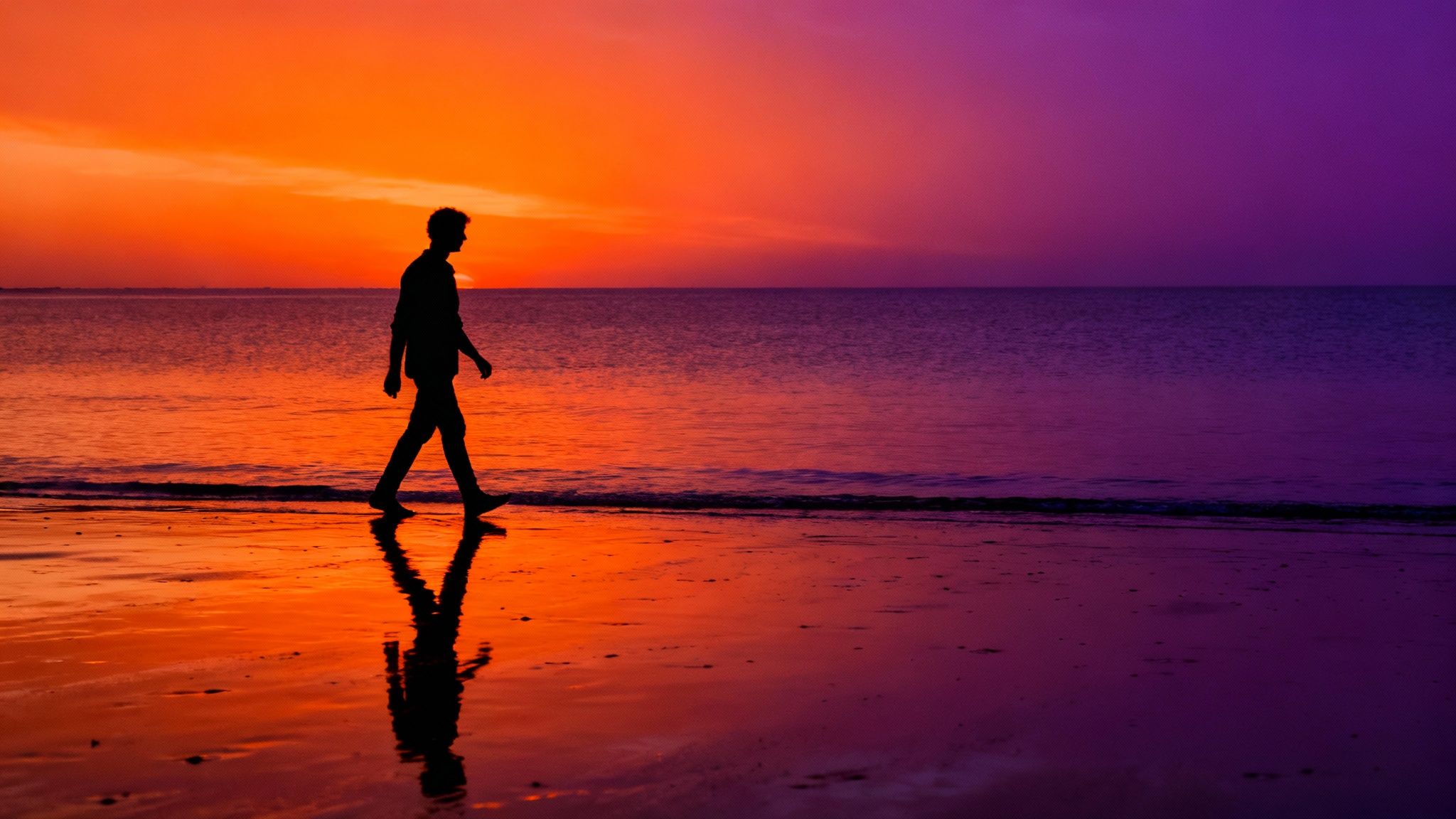 Silhouette of person walking along beach shore during vibrant orange and purple sunset reflection