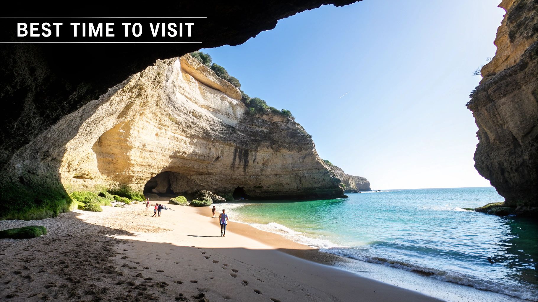 Sunny beach within a large sea cave with turquoise water, towering cliffs, and people walking.