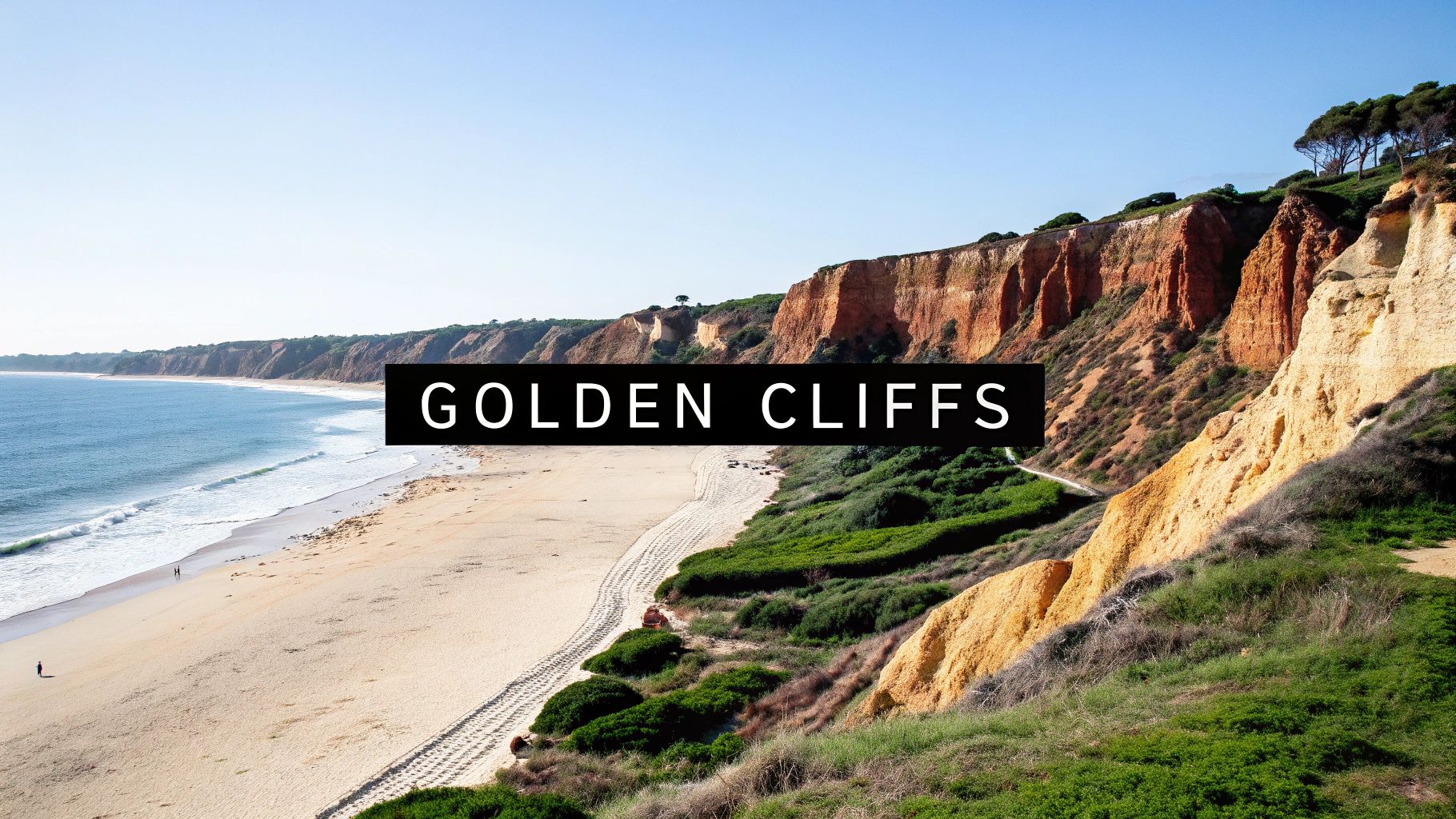 Scenic view of a long sandy beach, blue ocean, and towering golden cliffs under a clear sky.
