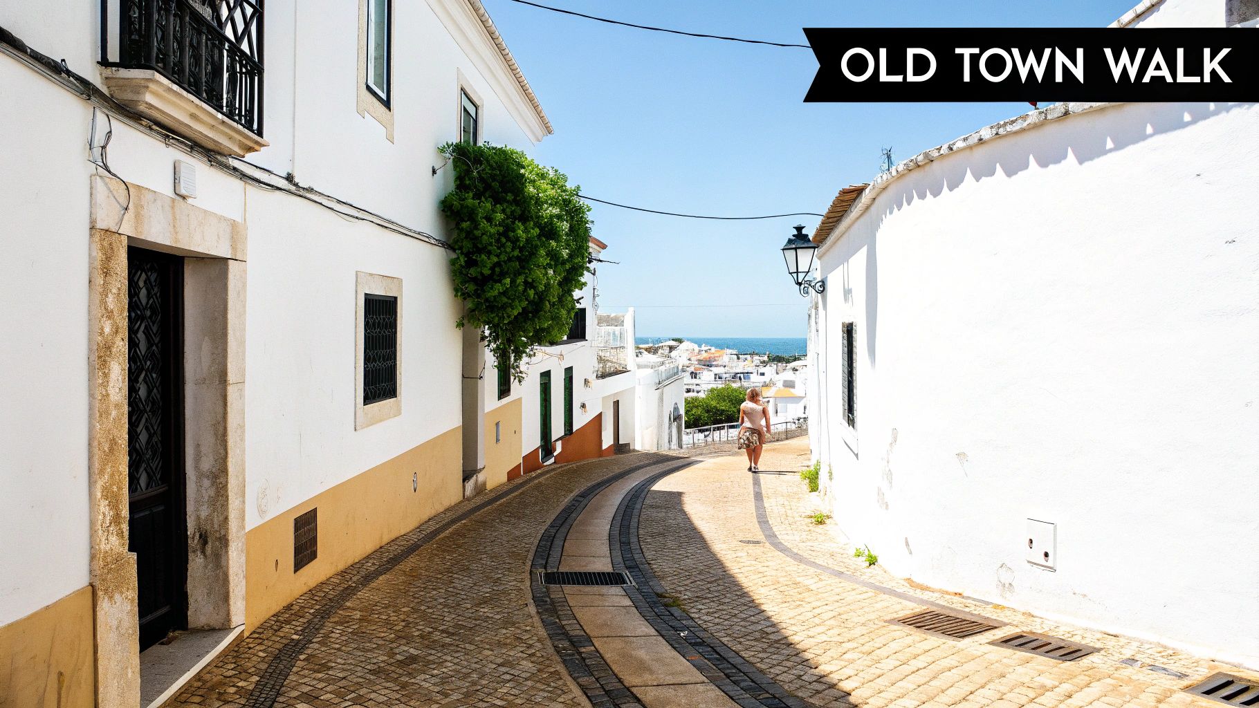 A person walks down a sunny, cobbled street in a historic white-walled old town with a distant sea view.