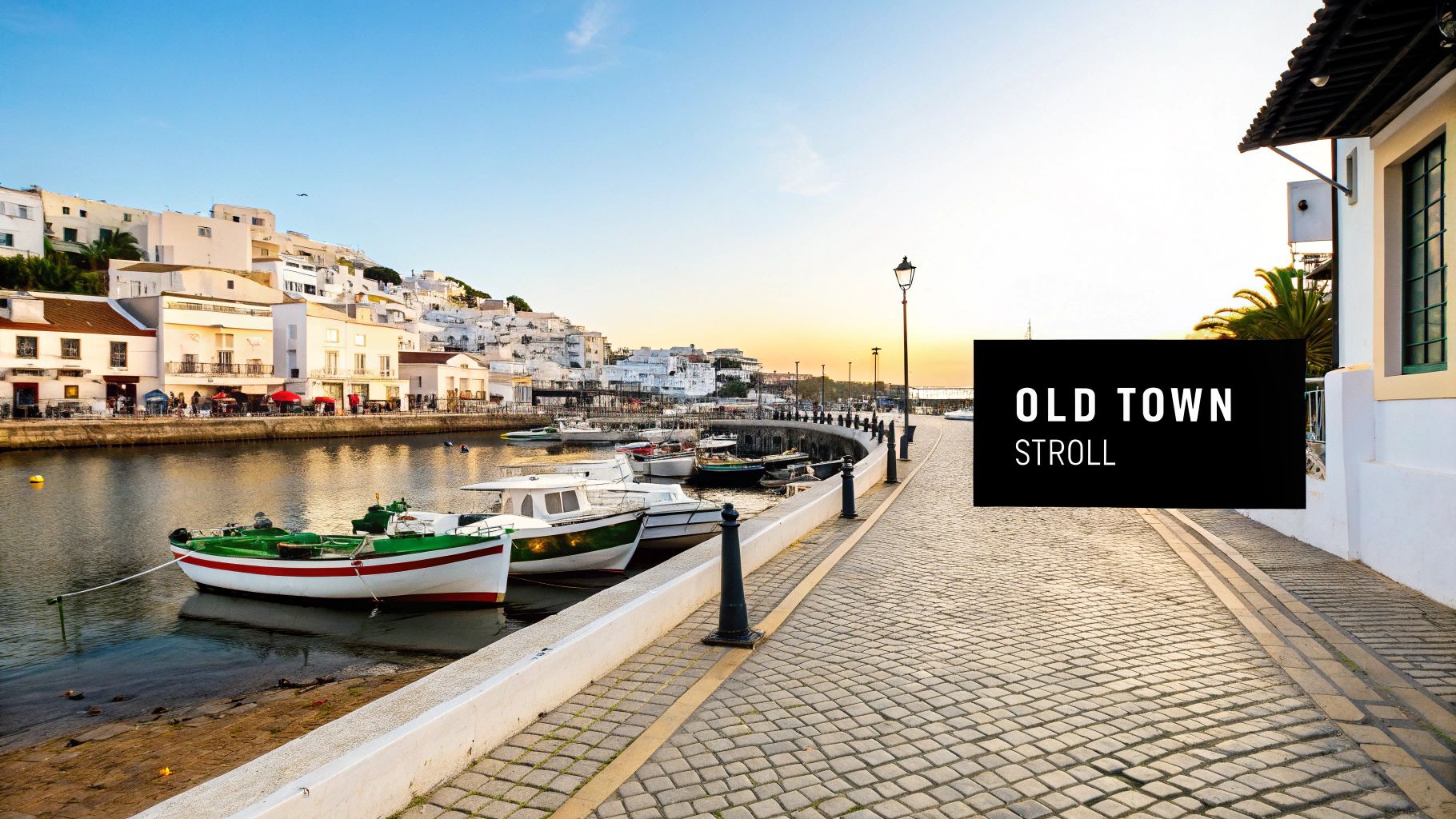 A picturesque old town waterfront with white buildings, colourful boats, and a cobblestone path at sunset.
