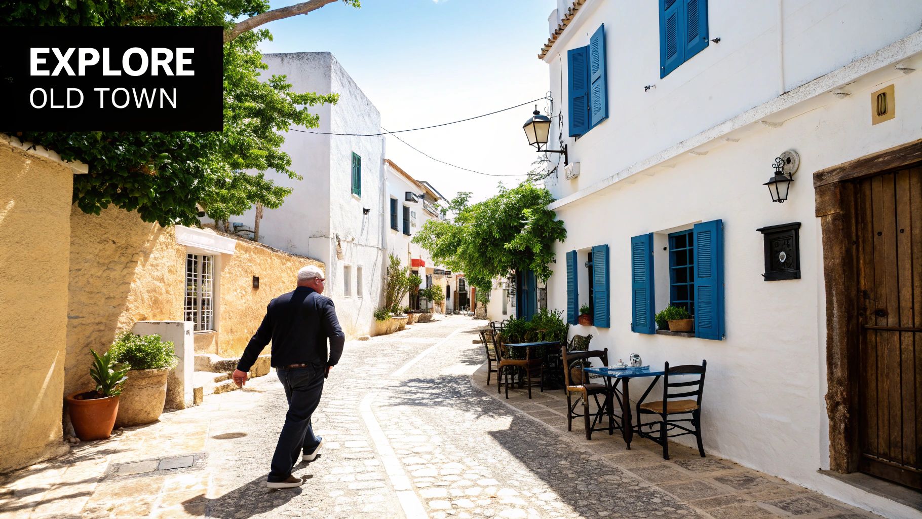 A man walks down a charming, sunlit narrow street in an old town with white buildings and blue shutters.