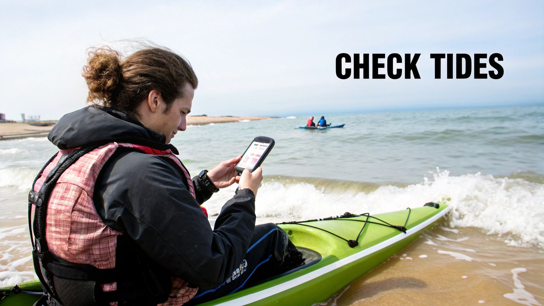 A kayaker on a beach checks their smartphone, possibly for tides, with another kayak in the background.