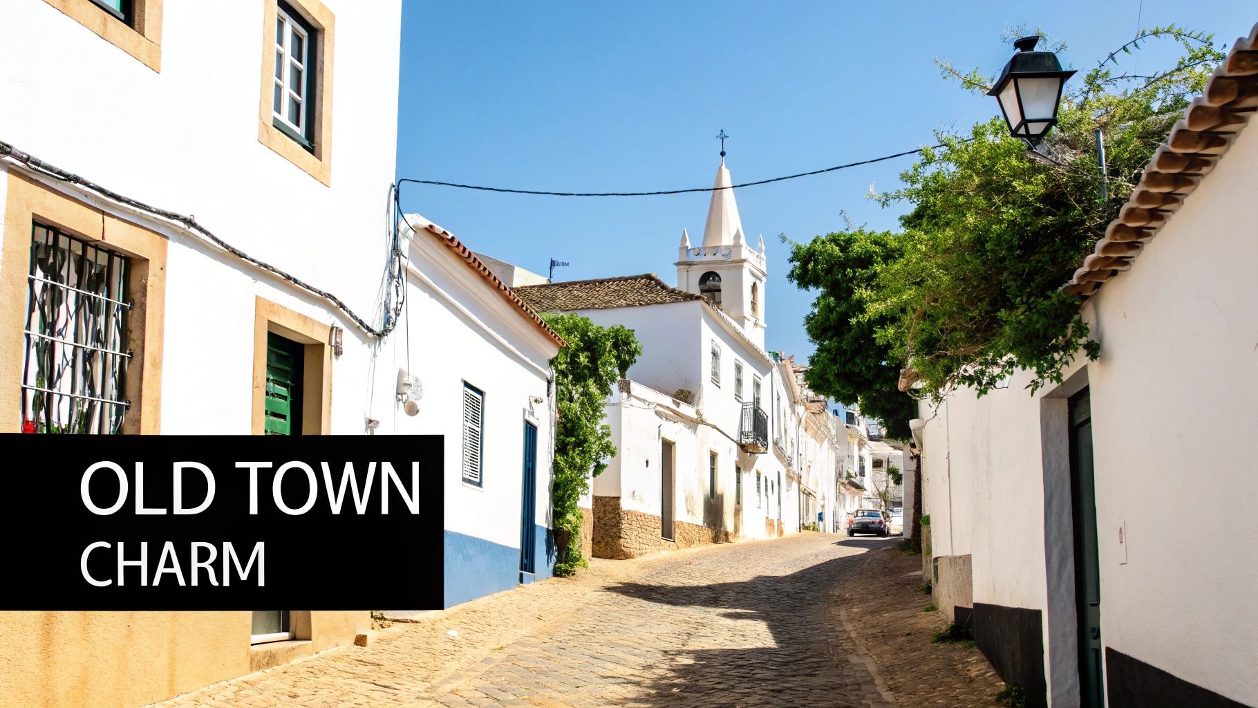 A picturesque, narrow cobblestone street winds through a charming old town with white buildings and a church steeple under a clear blue sky.