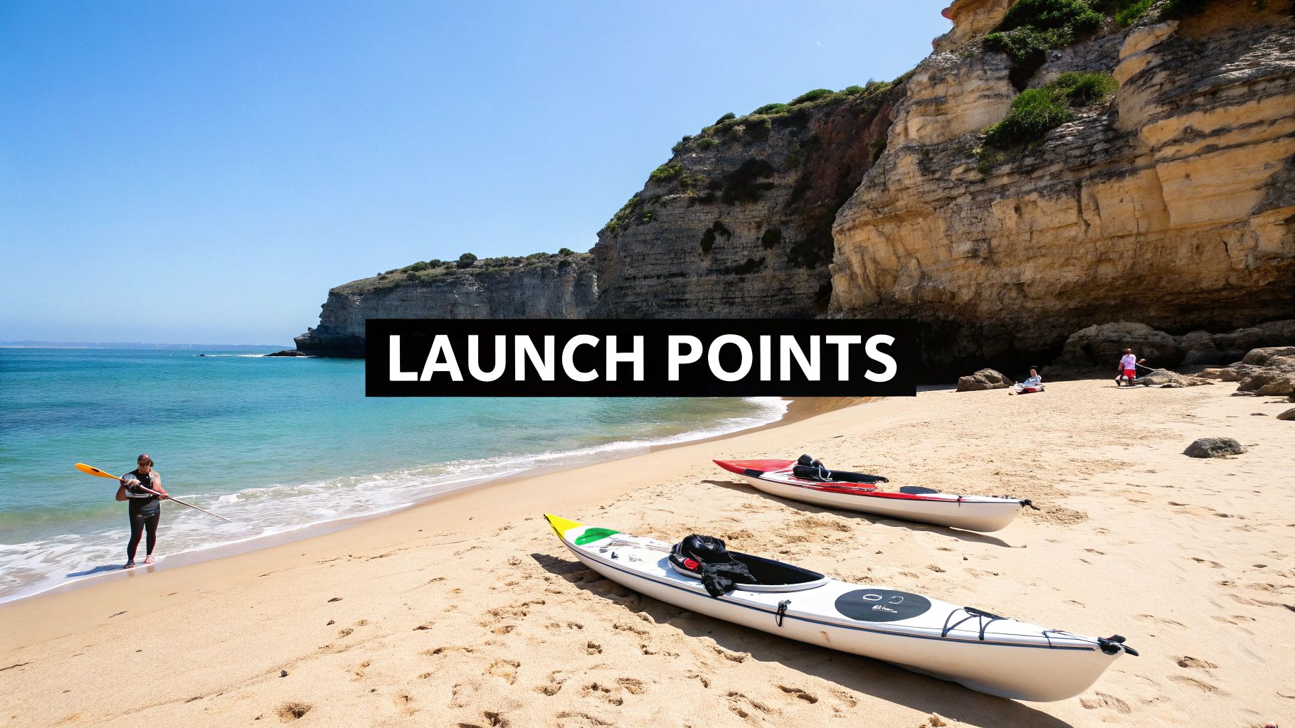 A person with a paddle stands in the water near two kayaks on a sandy beach with cliffs.