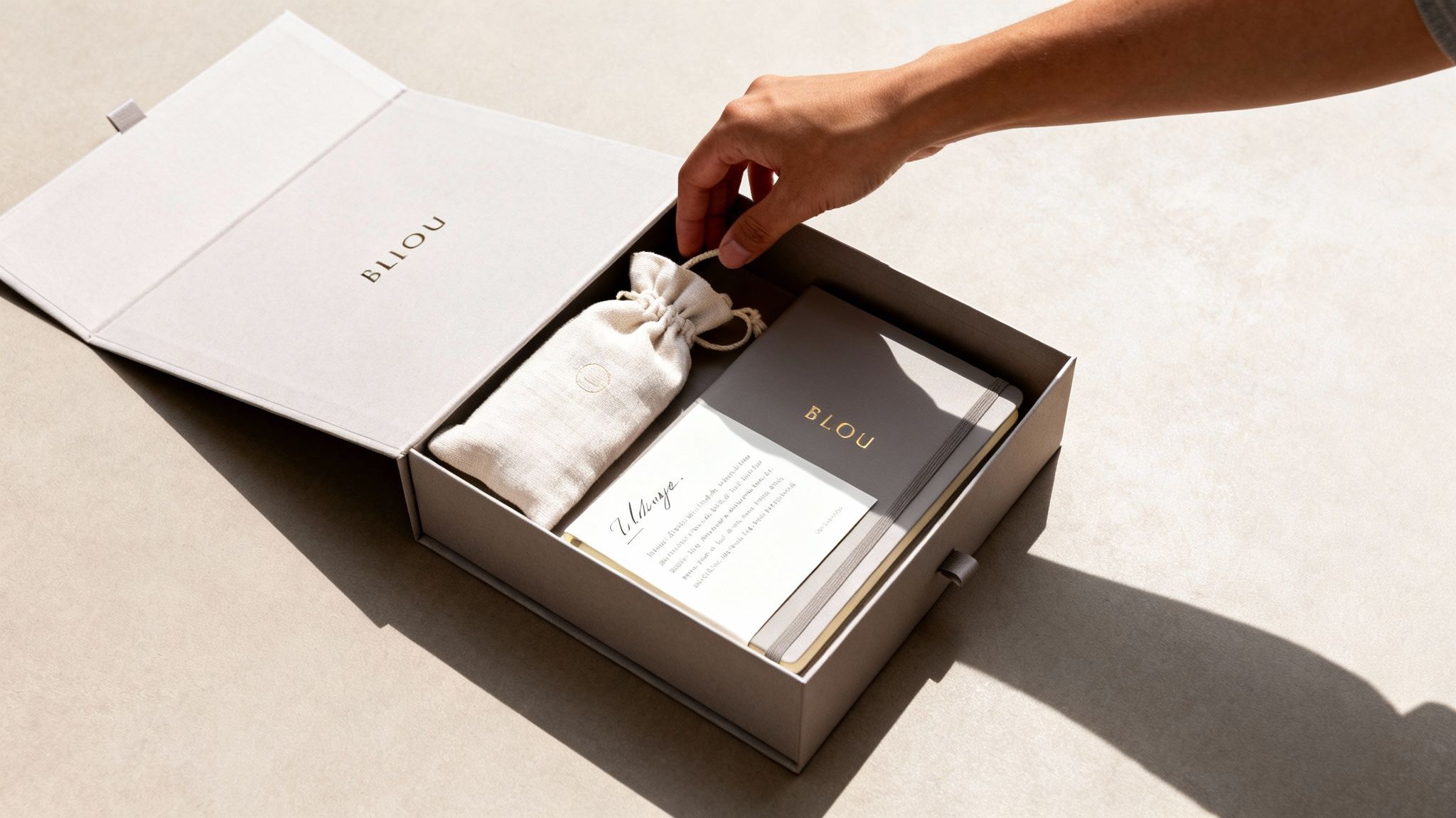 Eine Hand öffnet eine hellgraue Geschenkbox mit BLIOU-Branding, die einen Beutel, ein Notizbuch und eine Karte enthält.