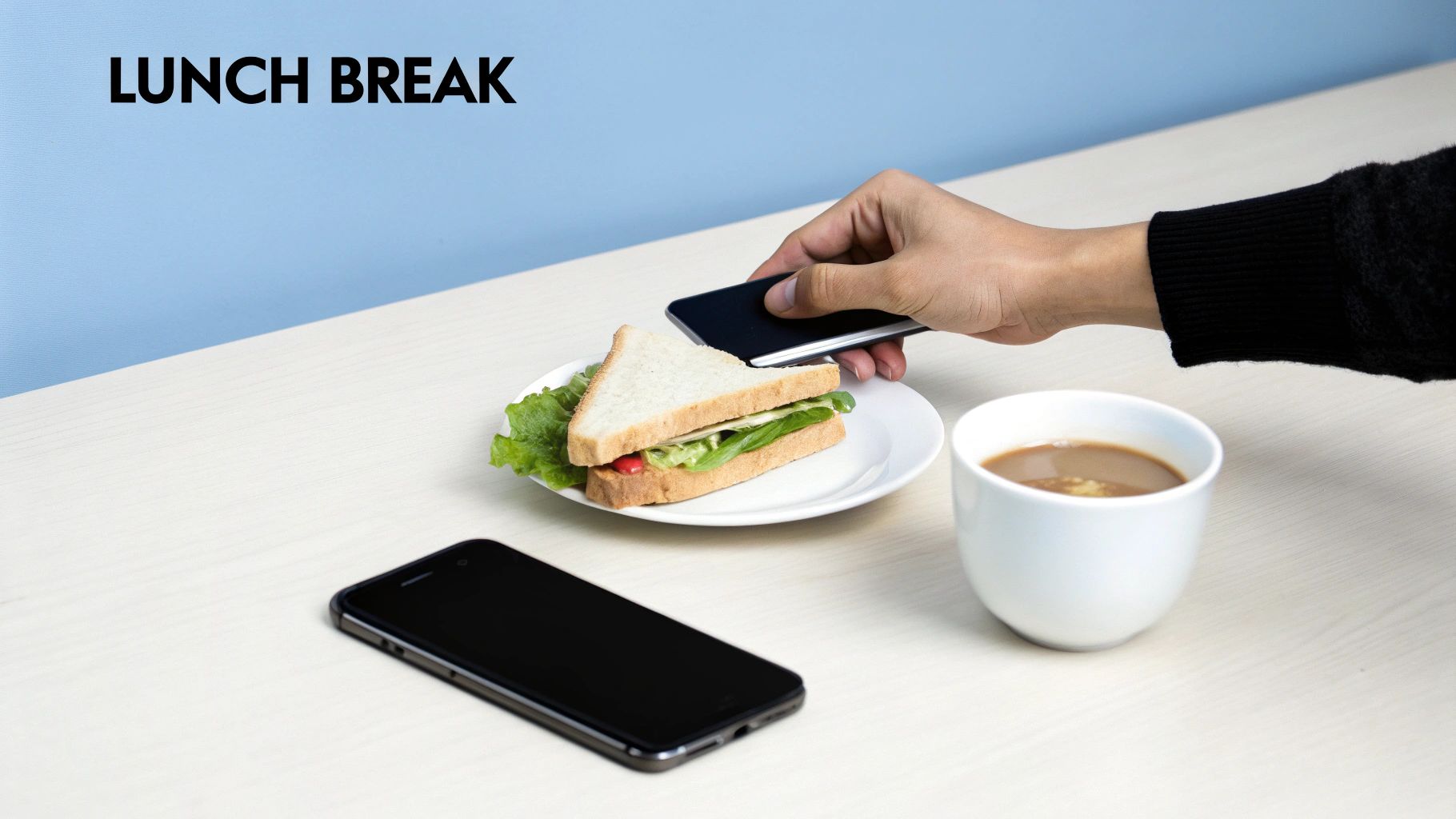 A hand holds a smartphone above a sandwich, coffee, and another phone on a table during a lunch break.