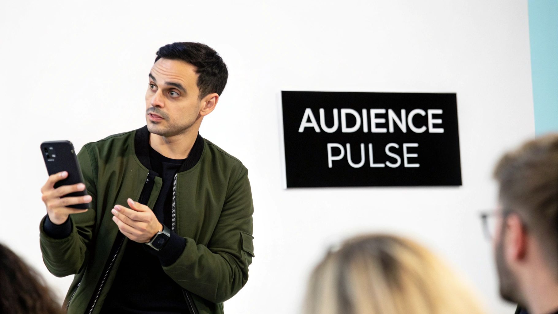 Man in green jacket holding a smartphone speaks to an audience under 'AUDIENCE PULSE' sign.