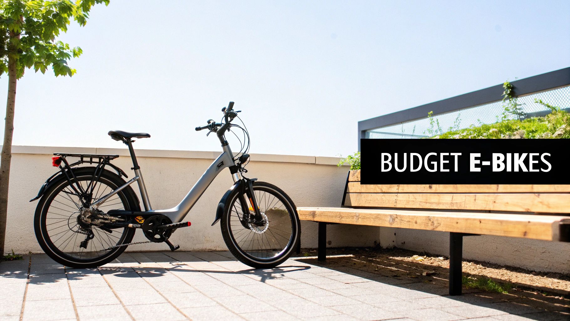 A grey electric bike is parked outdoors next to a wooden bench with a 'BUDGET E-BIKES' sign.