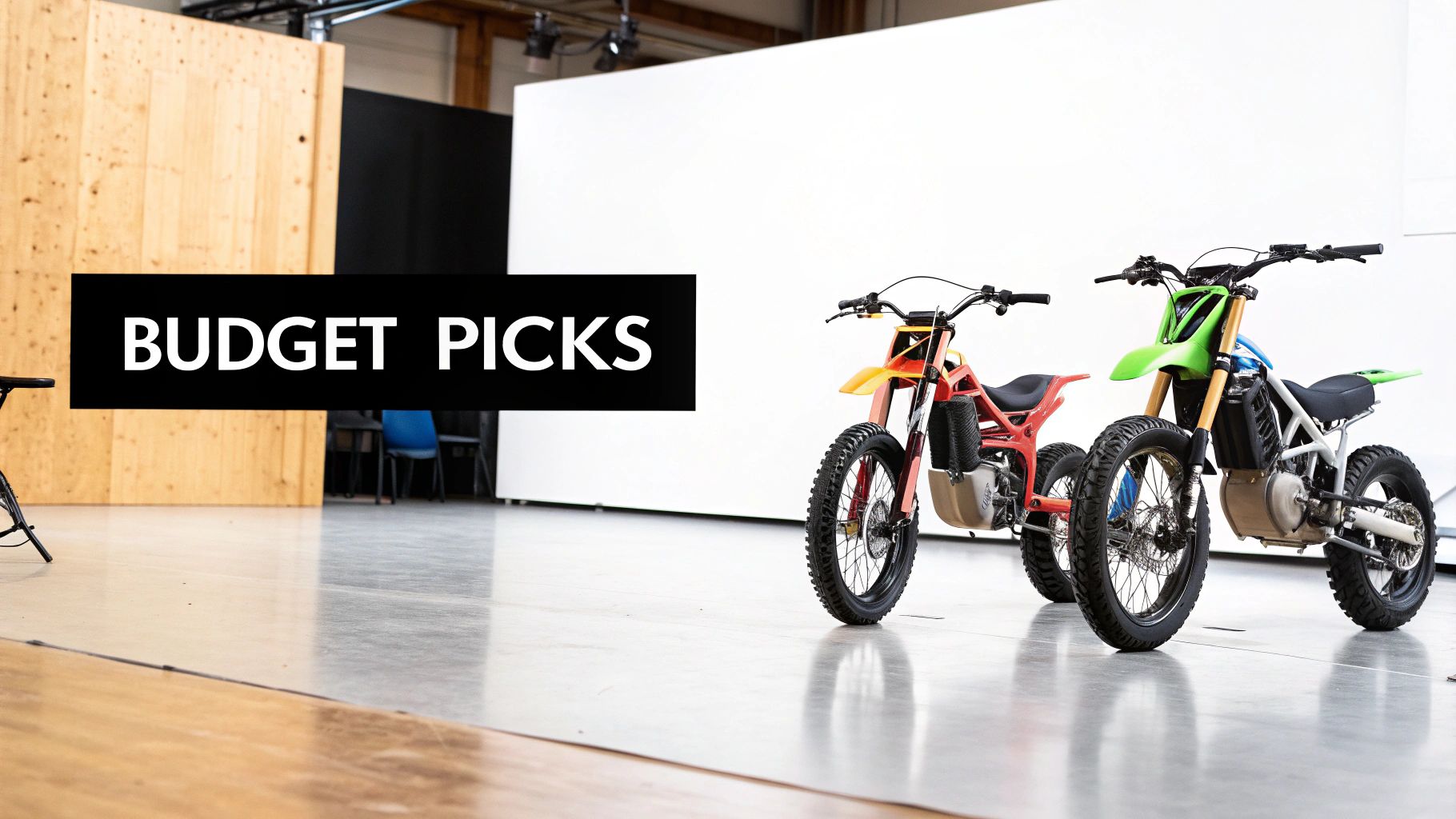 Two colorful electric dirt bikes, labeled "BUDGET PICKS", stand on a polished concrete floor in a studio.