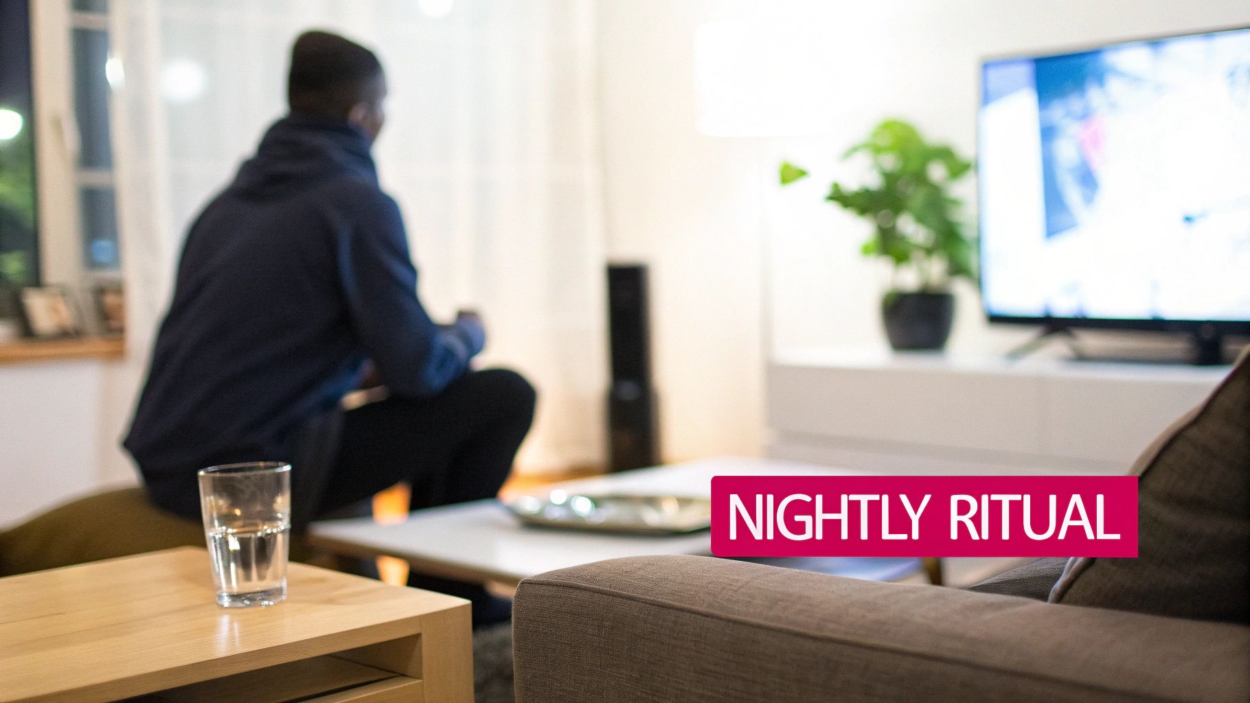 A person in a blue hoodie sits in a living room, focused on a TV screen at night.