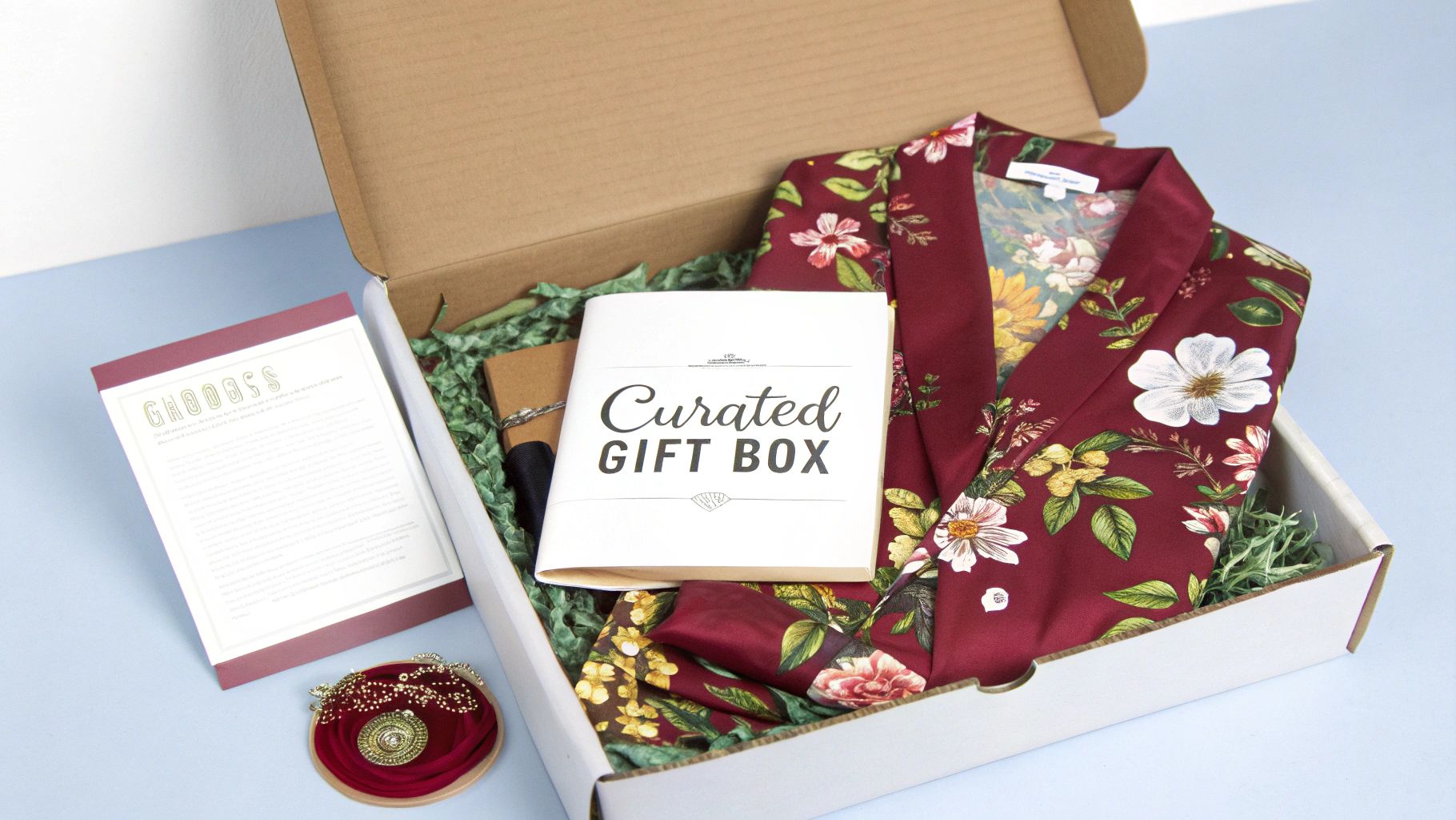 An open curated gift box filled with a burgundy floral robe, a booklet, and other thoughtful gifts.