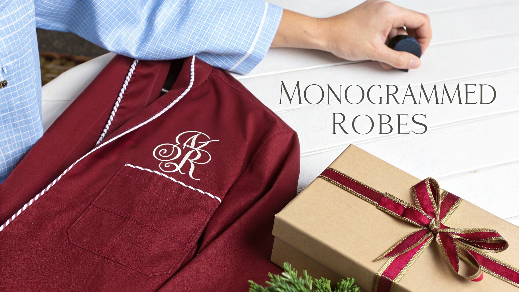 A burgundy monogrammed robe with white piping and 'AR' initials, next to a gift box with a red ribbon and a person's arm, with the text 'MONOGRAMMED ROBES'.