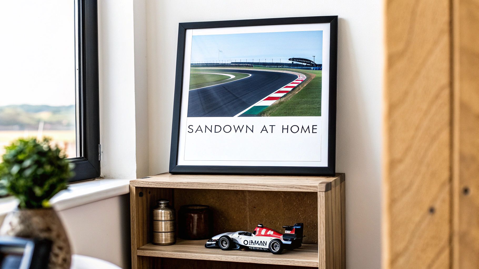 A framed picture of a race track with 'Sandown at Home' text on a shelf with a toy race car.