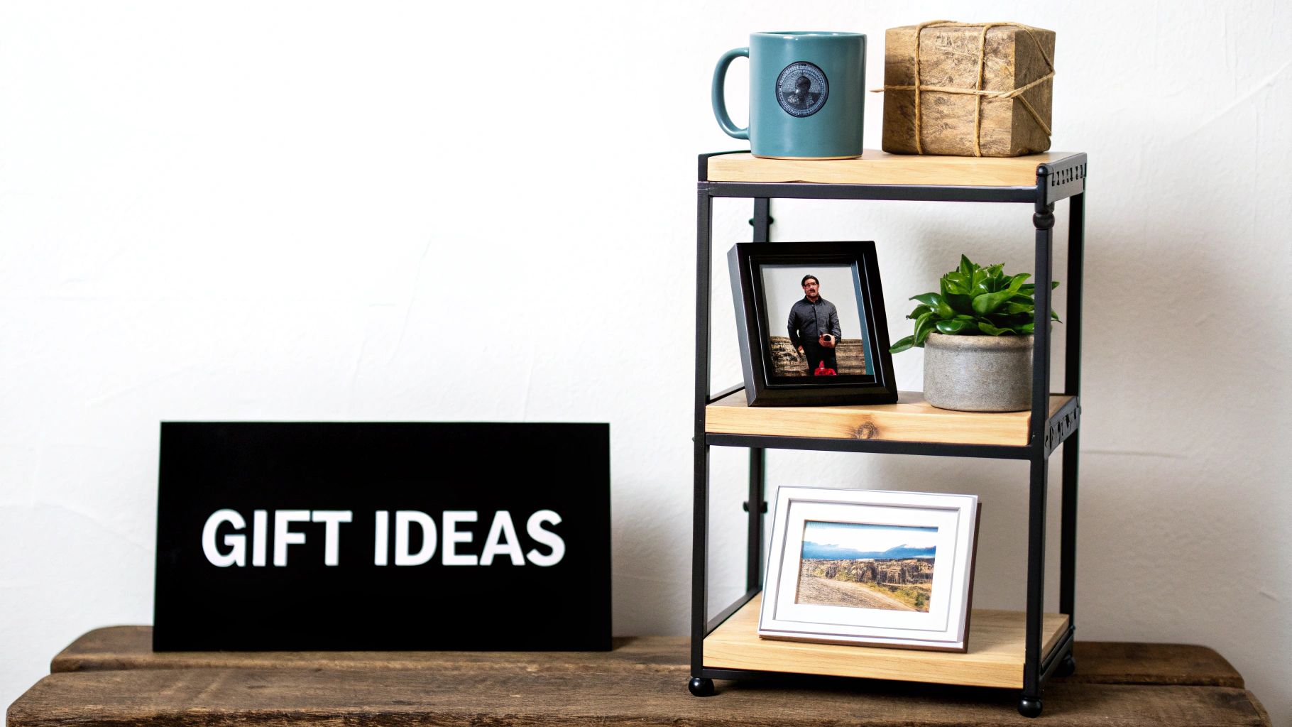 A black sign reading 'GIFT IDEAS' next to a wooden shelf displaying a mug, wrapped gift, framed photos, and a potted plant.