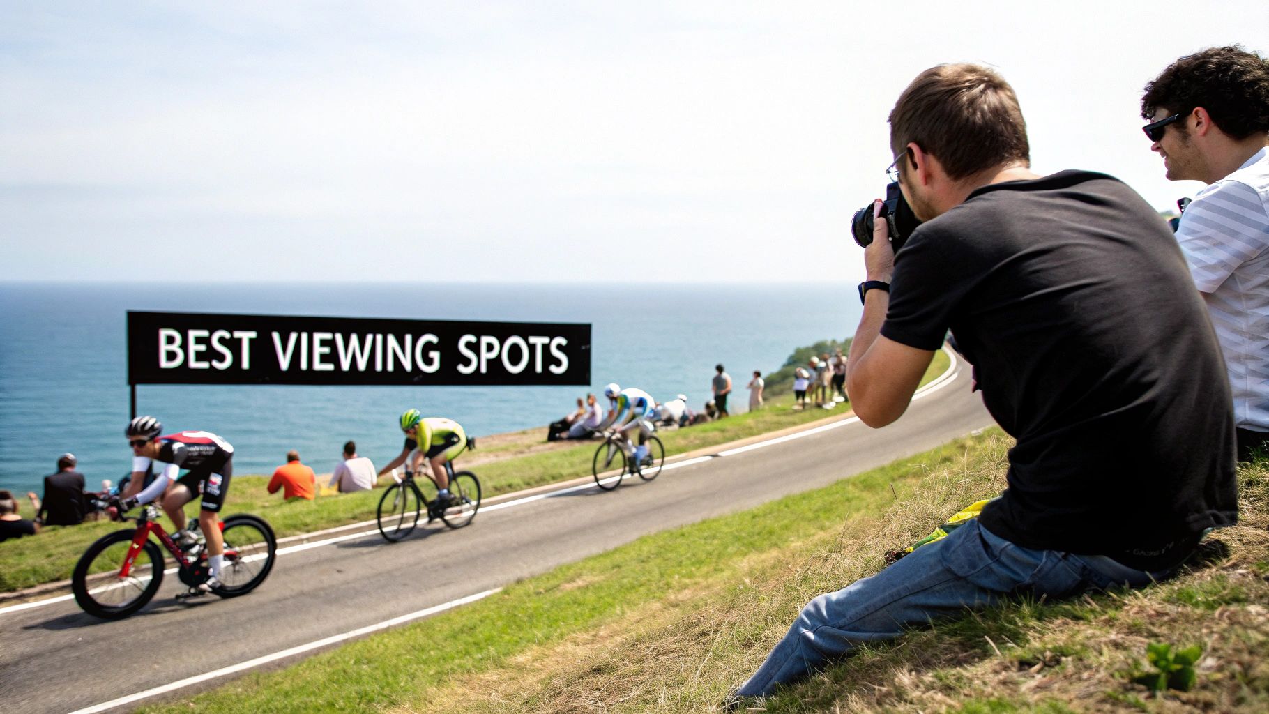 Spectators and a photographer capturing cyclists racing along a scenic coastal road with a "BEST VIEWING SPOTS" sign.