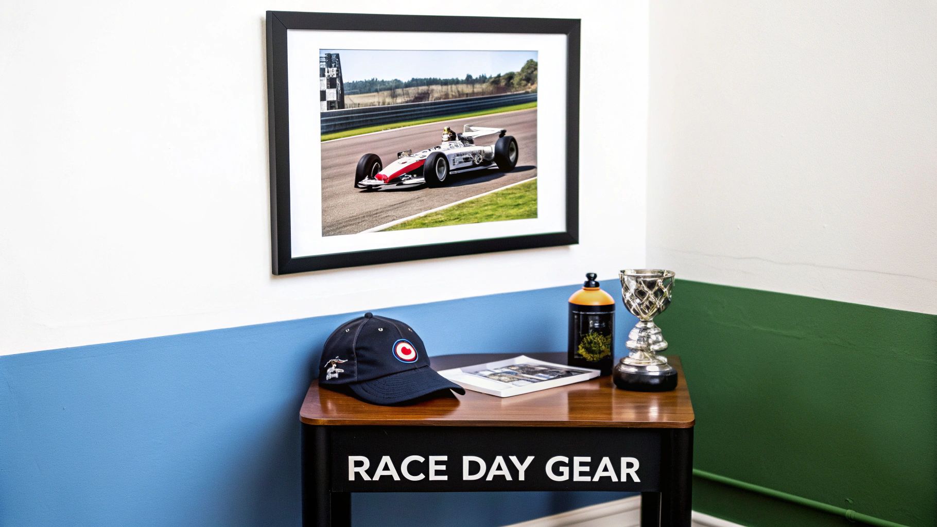 A framed photo of a race car on a track hangs above a table with race day gear including a cap, trophy, and book.