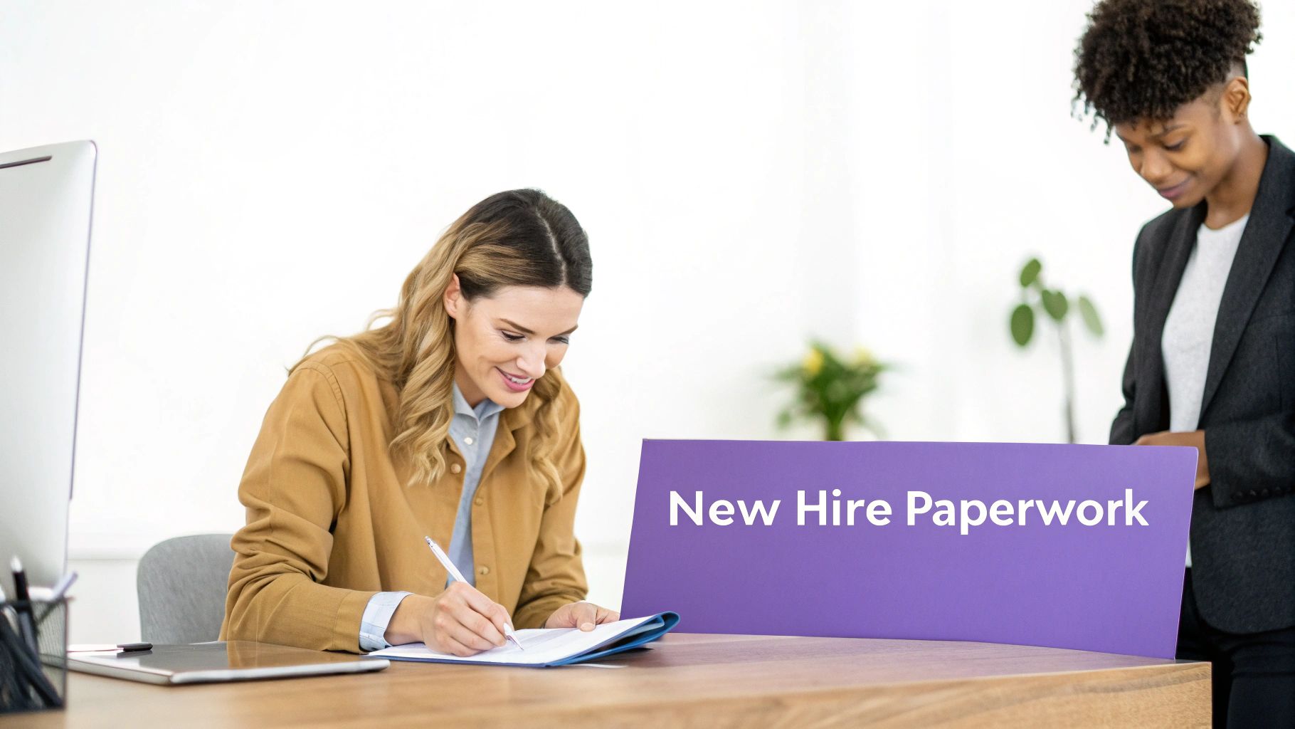 A smiling woman signs new hire paperwork at a desk, with another woman standing nearby.