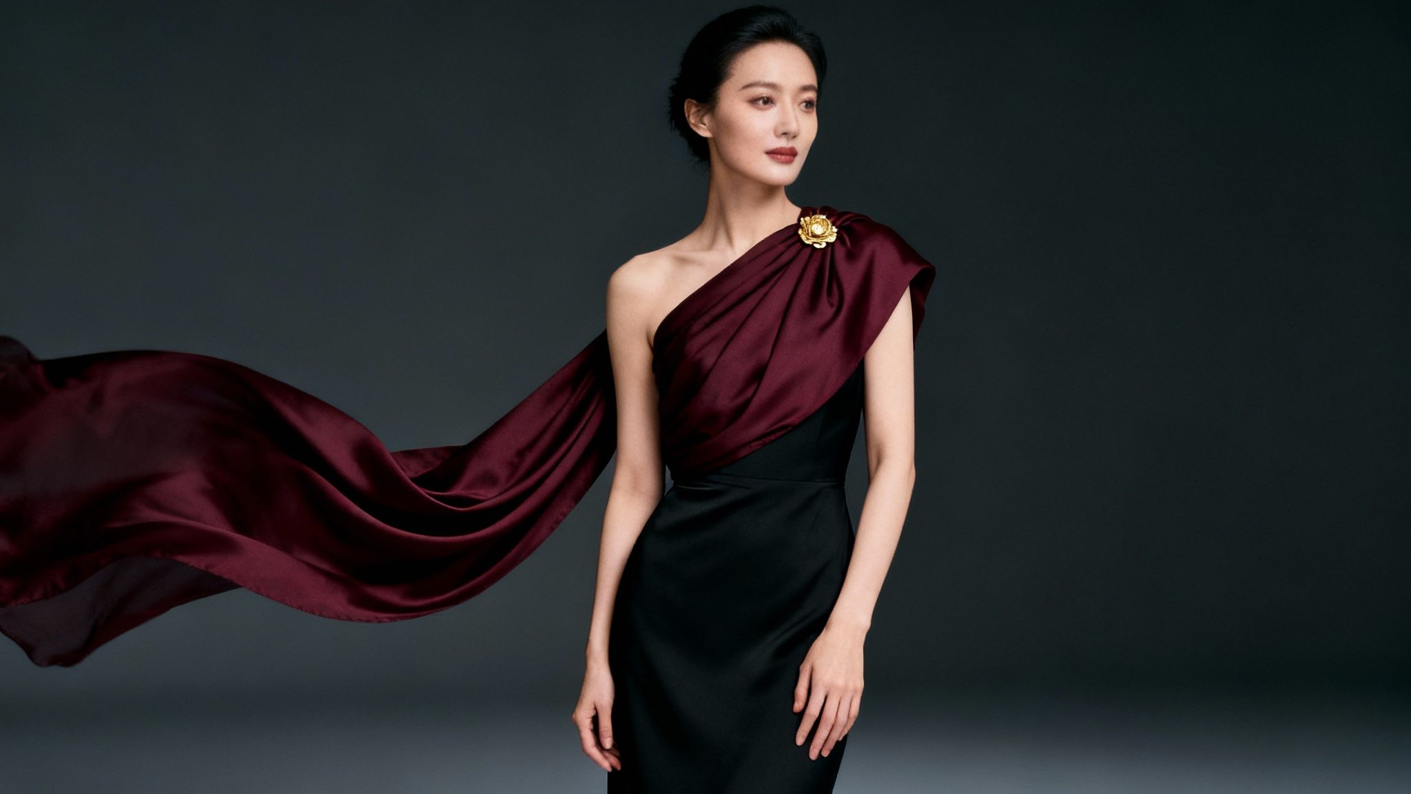 Elegant woman in a black and burgundy one-shoulder dress with a flowing scarf and golden brooch.