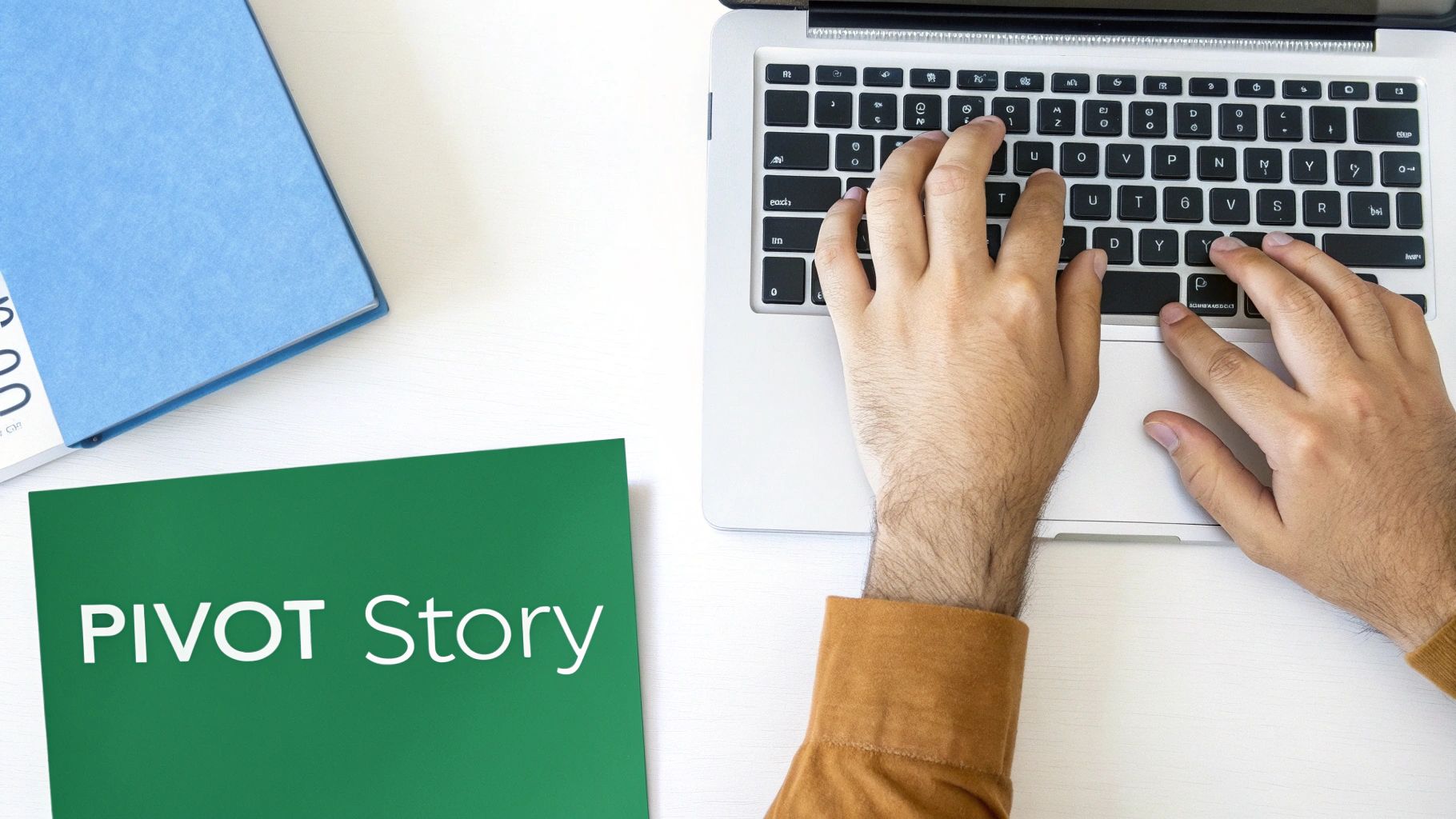 Overhead shot of hands typing on a laptop, with a "PIVOT Story" folder and blue book on a white desk.
