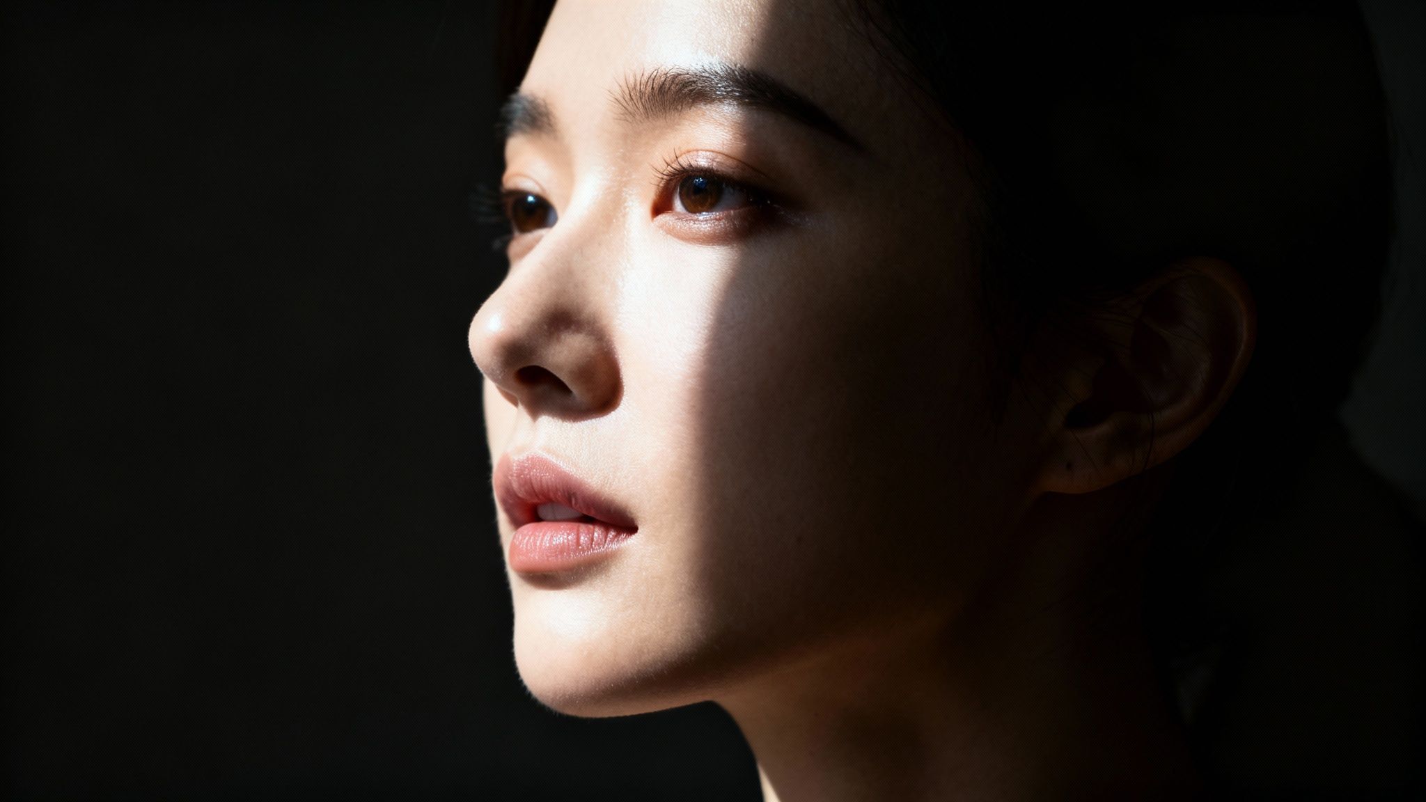 A young woman's face in profile, illuminated by a beam of light creating dramatic contrast against a dark background.