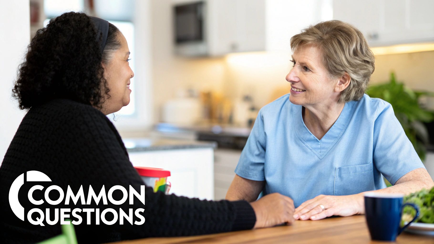 Two smiling women, one in blue scrubs, engaging in a friendly conversation in a kitchen.