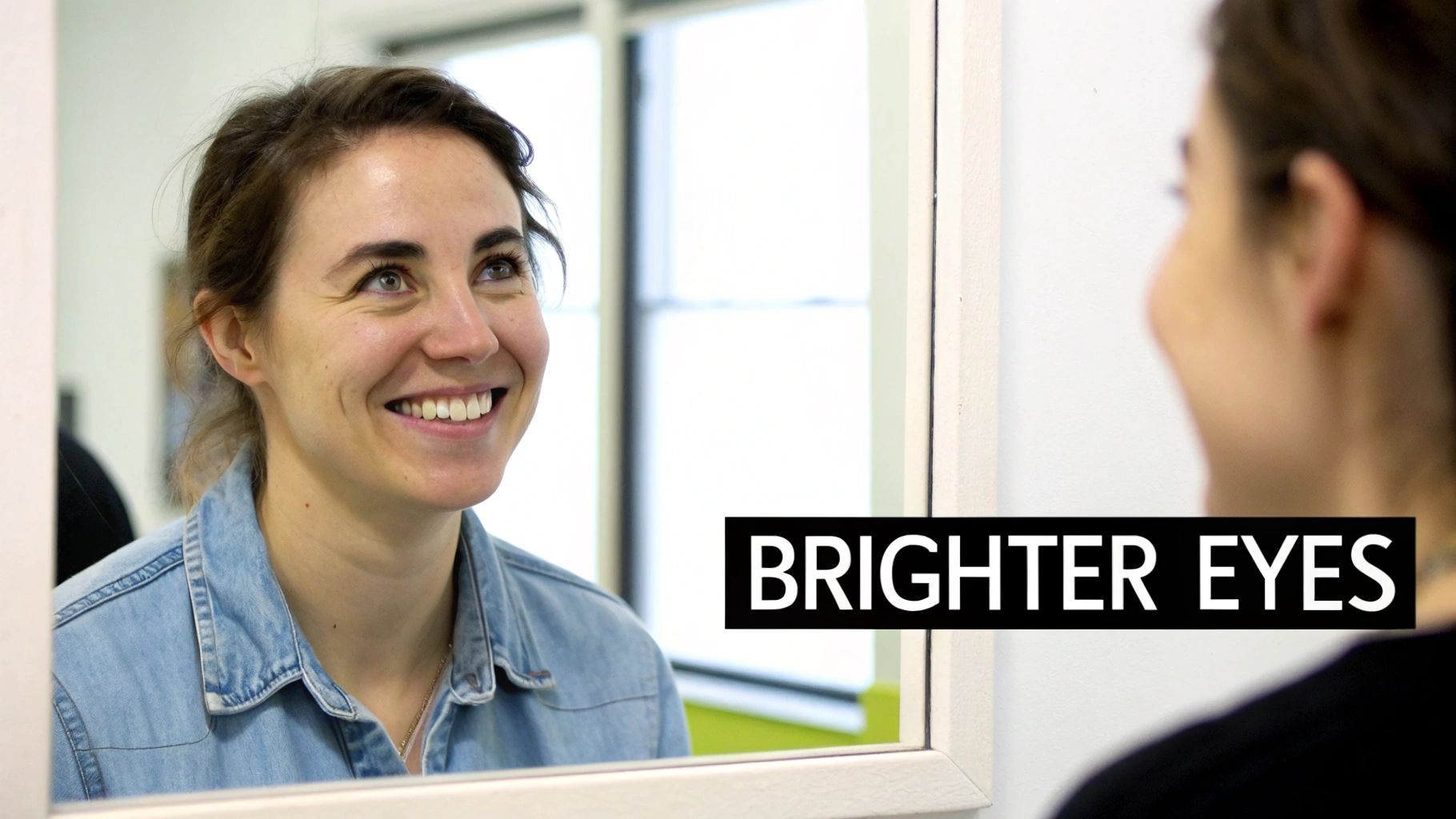 A smiling woman with bright eyes looks at her reflection in a mirror, wearing a denim shirt.