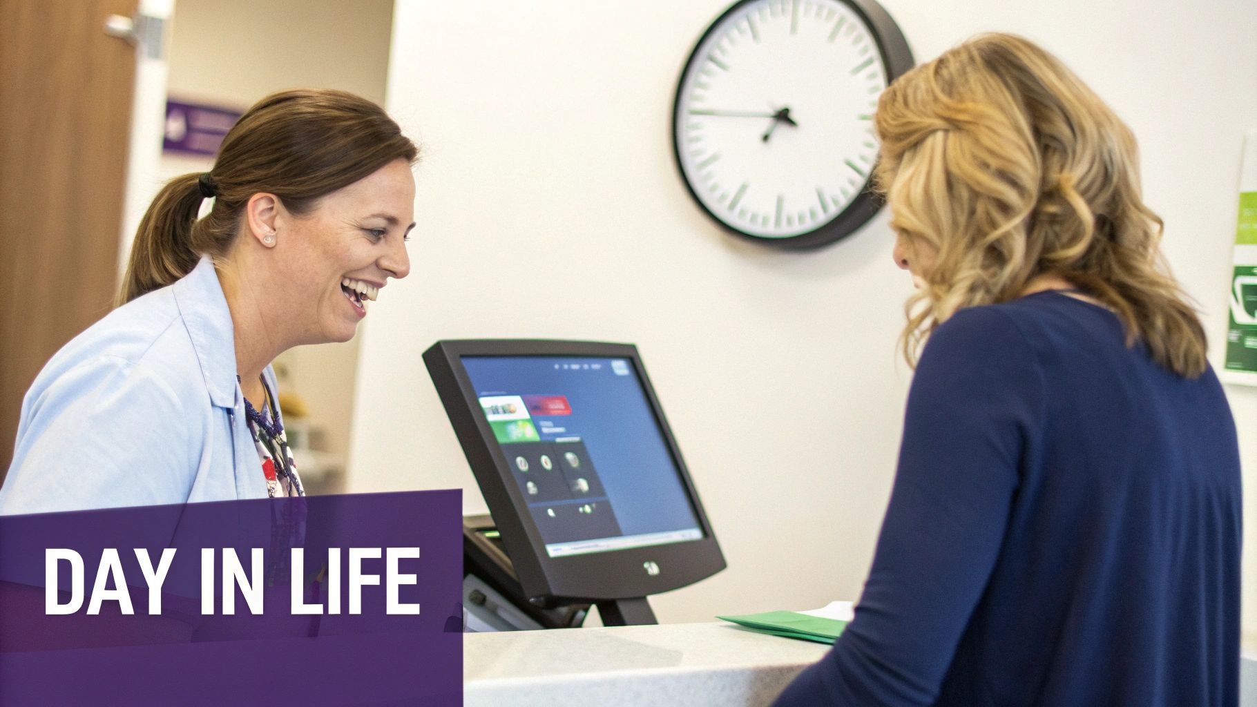A smiling medical receptionist helps a patient at a front desk with a computer screen nearby.