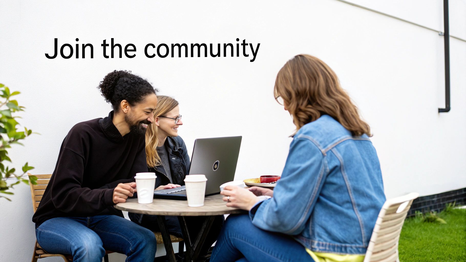 Three diverse people collaborating outdoors at a wooden table with a laptop, smiling and having coffee.