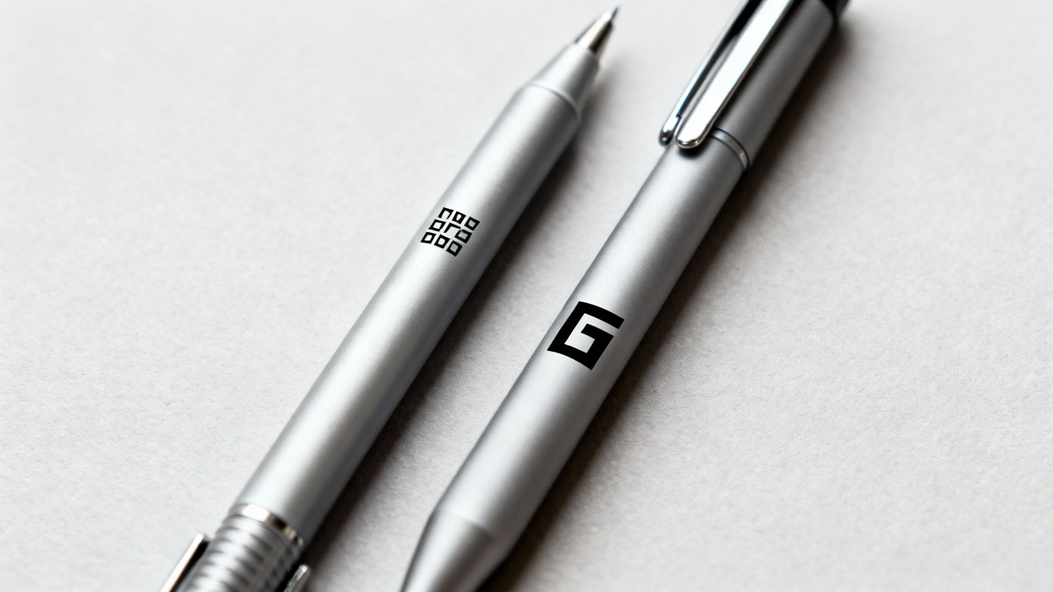 Close-up of two silver pens, one with a square grid logo and the other with a stylized G logo.