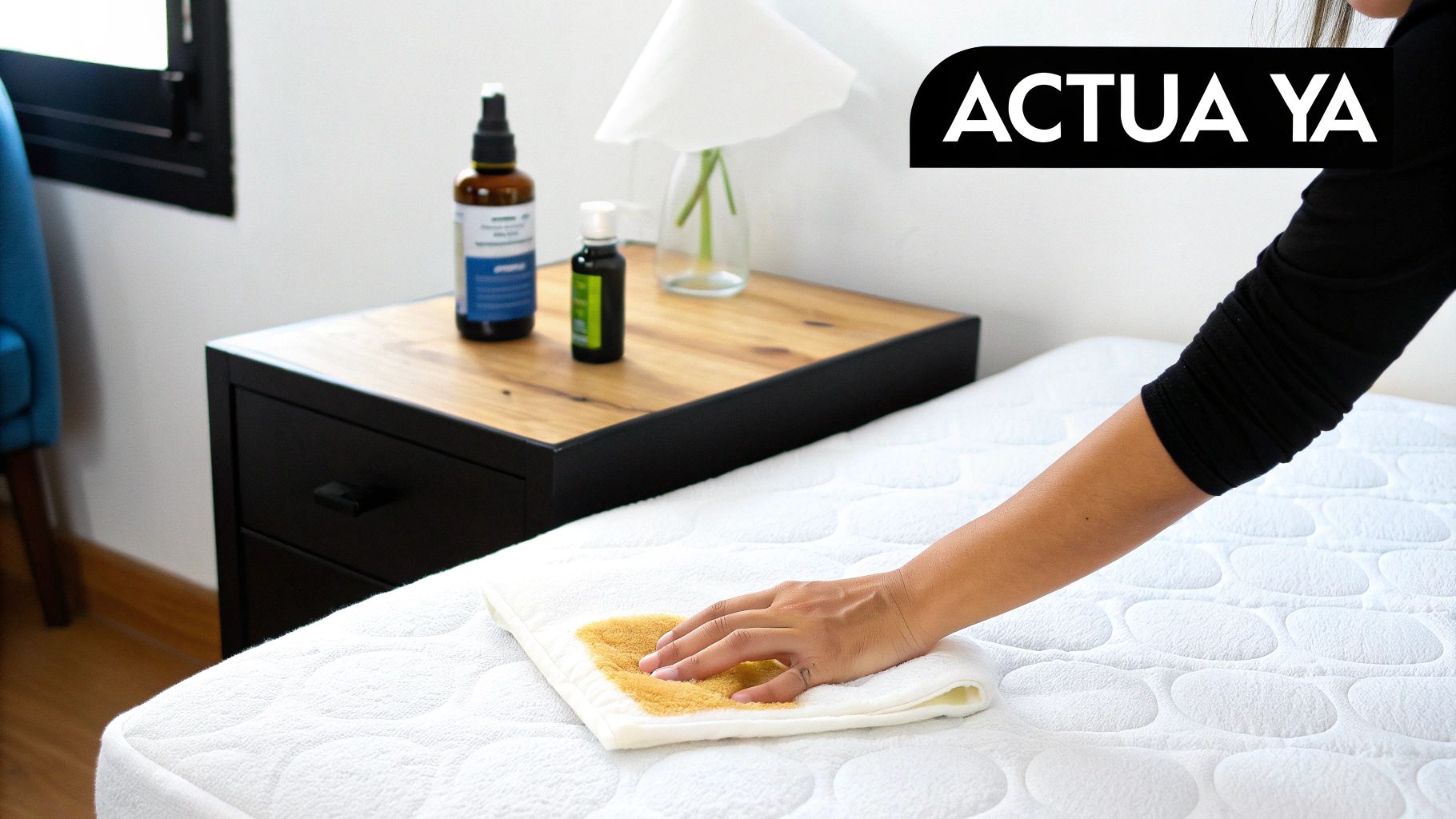 Persona limpiando una mancha amarilla de orina en un colchón blanco con un paño, junto a productos de limpieza.